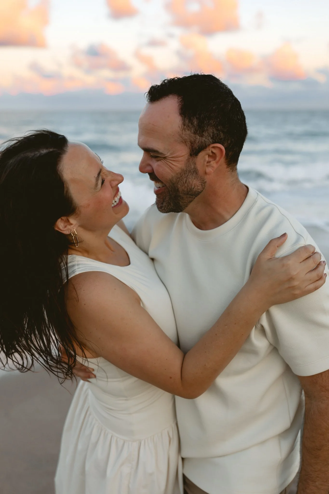 A happy couple embracing on the beach with ocean waves and a sunset sky in the background.