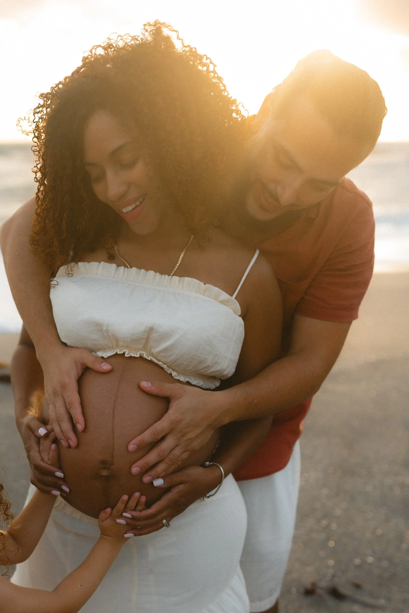 A happy pregnant woman on the beach with her partner and a child, all smiling and touching her belly.