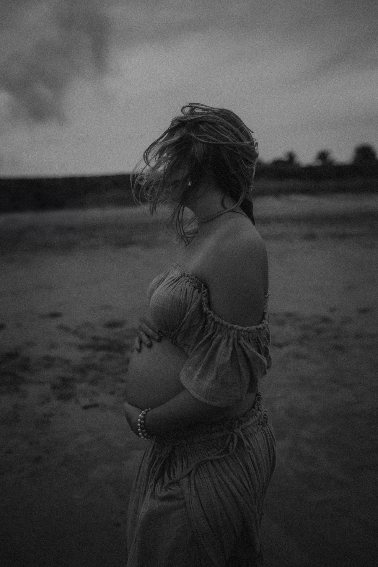 A pregnant woman with her hair blowing in the wind, standing on a beach at dusk or dawn, wearing a loose off-shoulder top and pants, holding her belly with one hand.