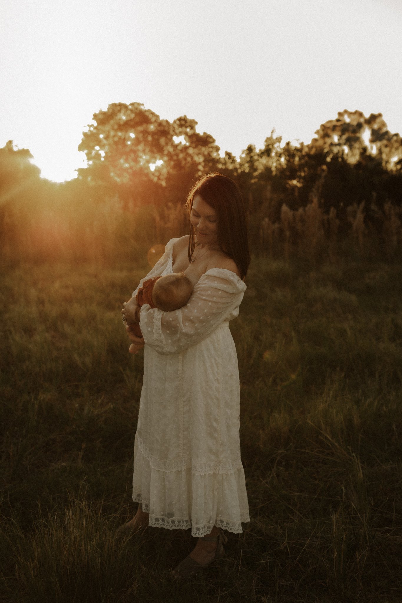 A woman in a long white dress holding a baby outdoors during sunset with trees in the background.