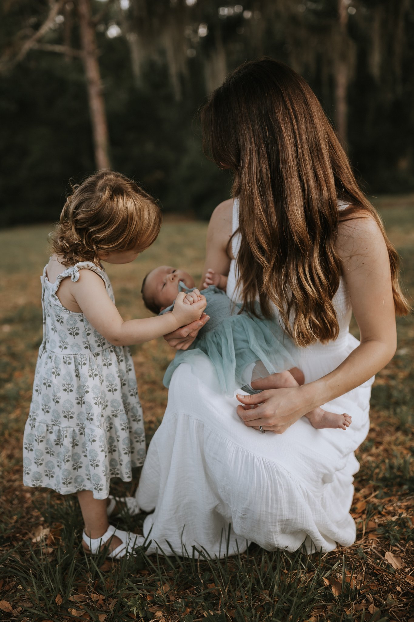 A woman holding a newborn baby outdoors, with a young girl touching the baby's hand, in a natural setting with trees and fallen leaves.