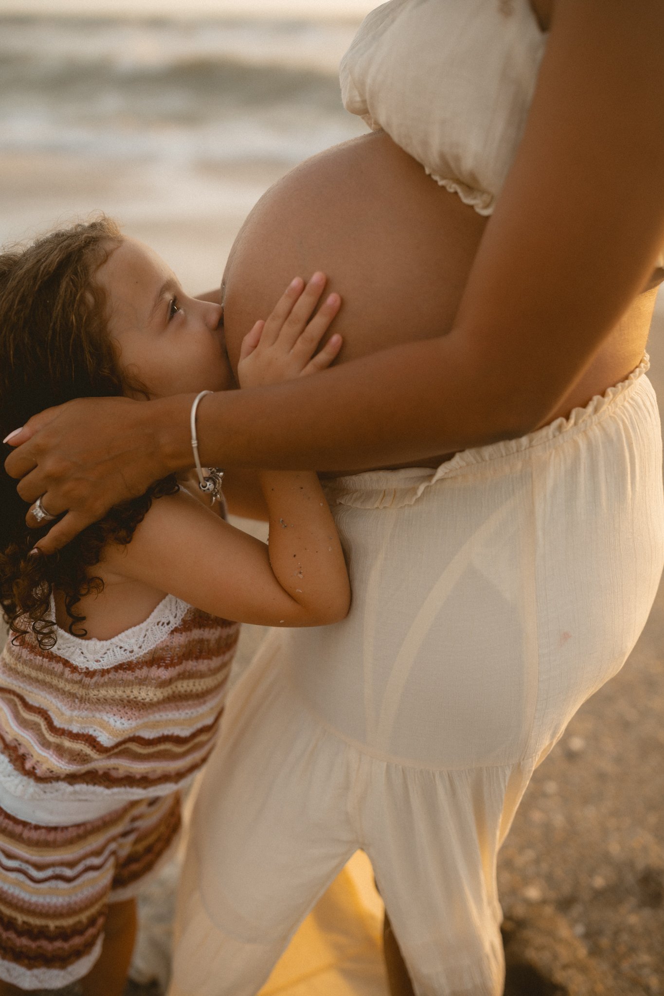 A young girl with dark curly hair kisses a pregnant woman's belly on the beach, with the ocean in the background.