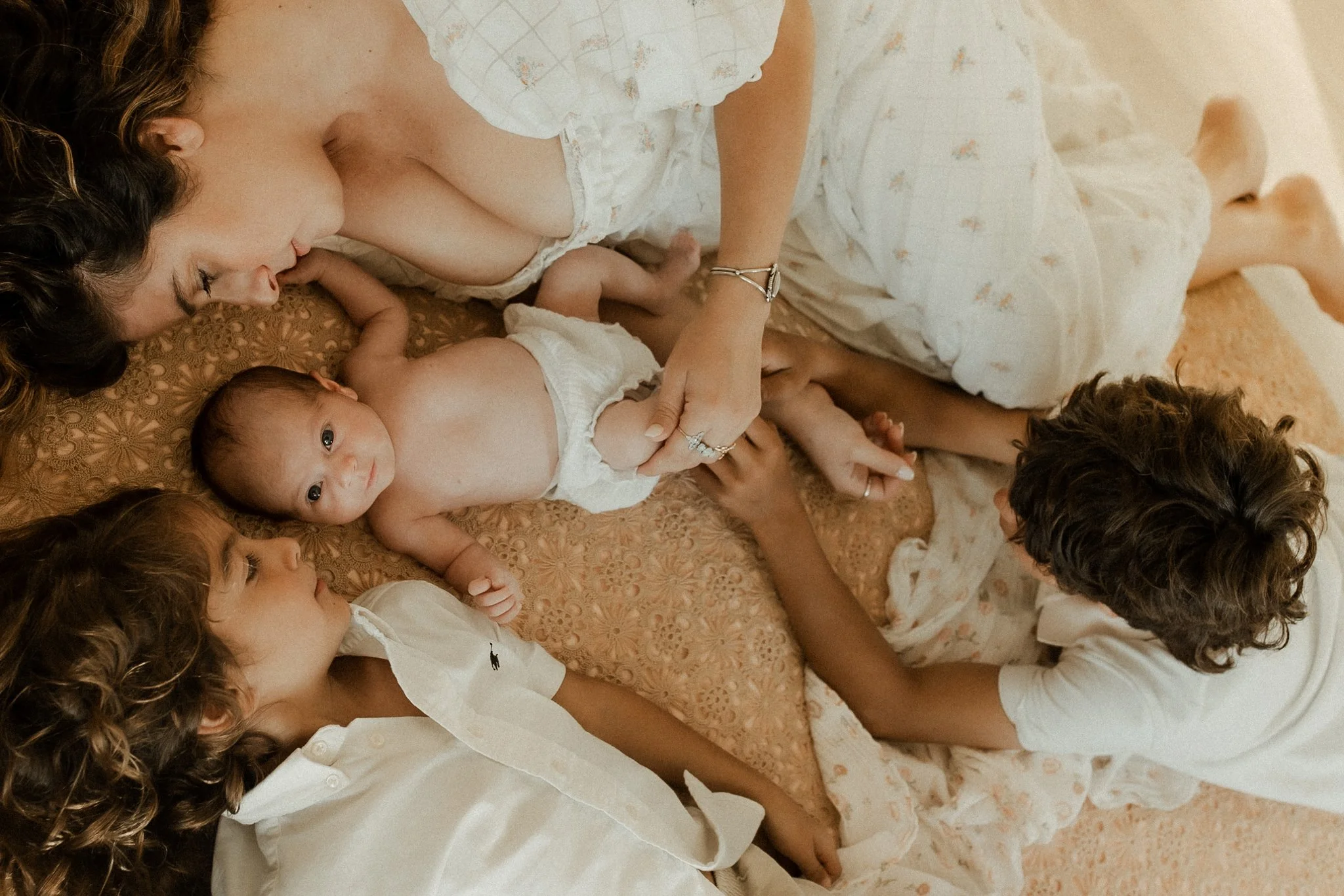 A woman lying on a bed with her three children, two girls and a baby boy, all in light-colored clothing, engaging in a tender moment.
