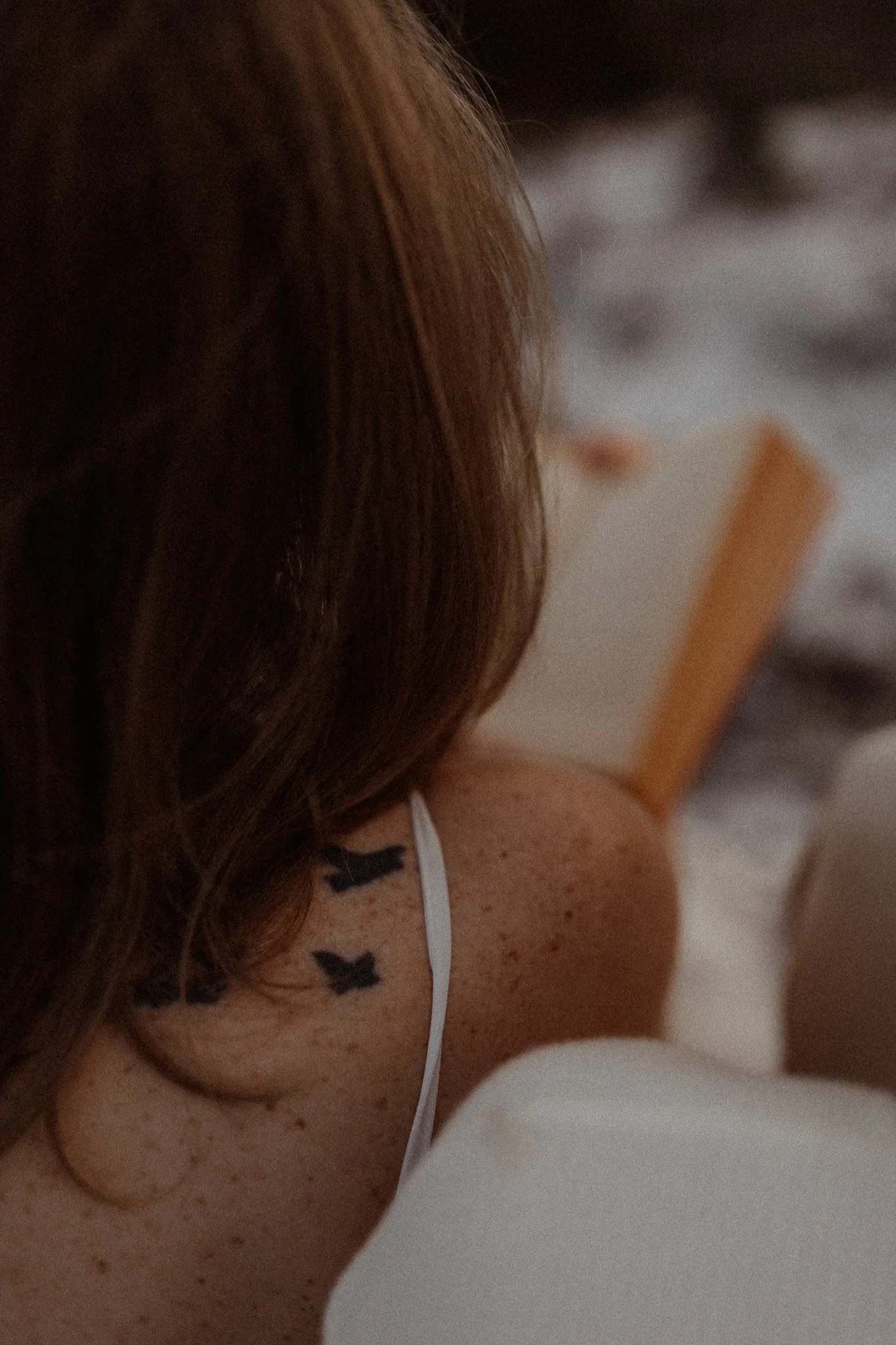 Close-up of a woman's shoulder and hair, showing small black butterfly tattoos on her shoulder, with a white strap of clothing visible and a blurred background.