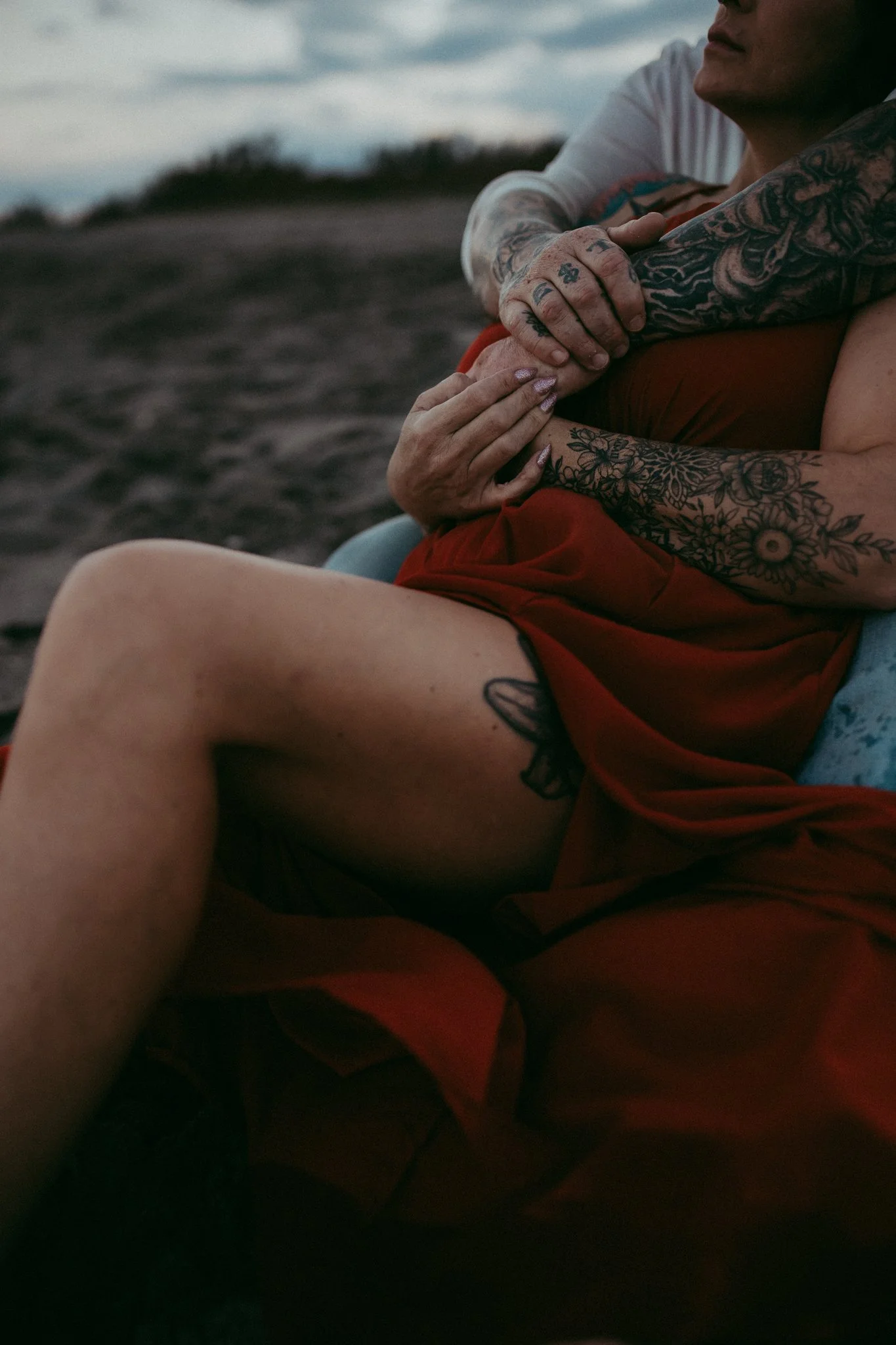 Two people sitting outdoors, embracing each other. One person's arms are tattoos, and both are wearing casual clothing.