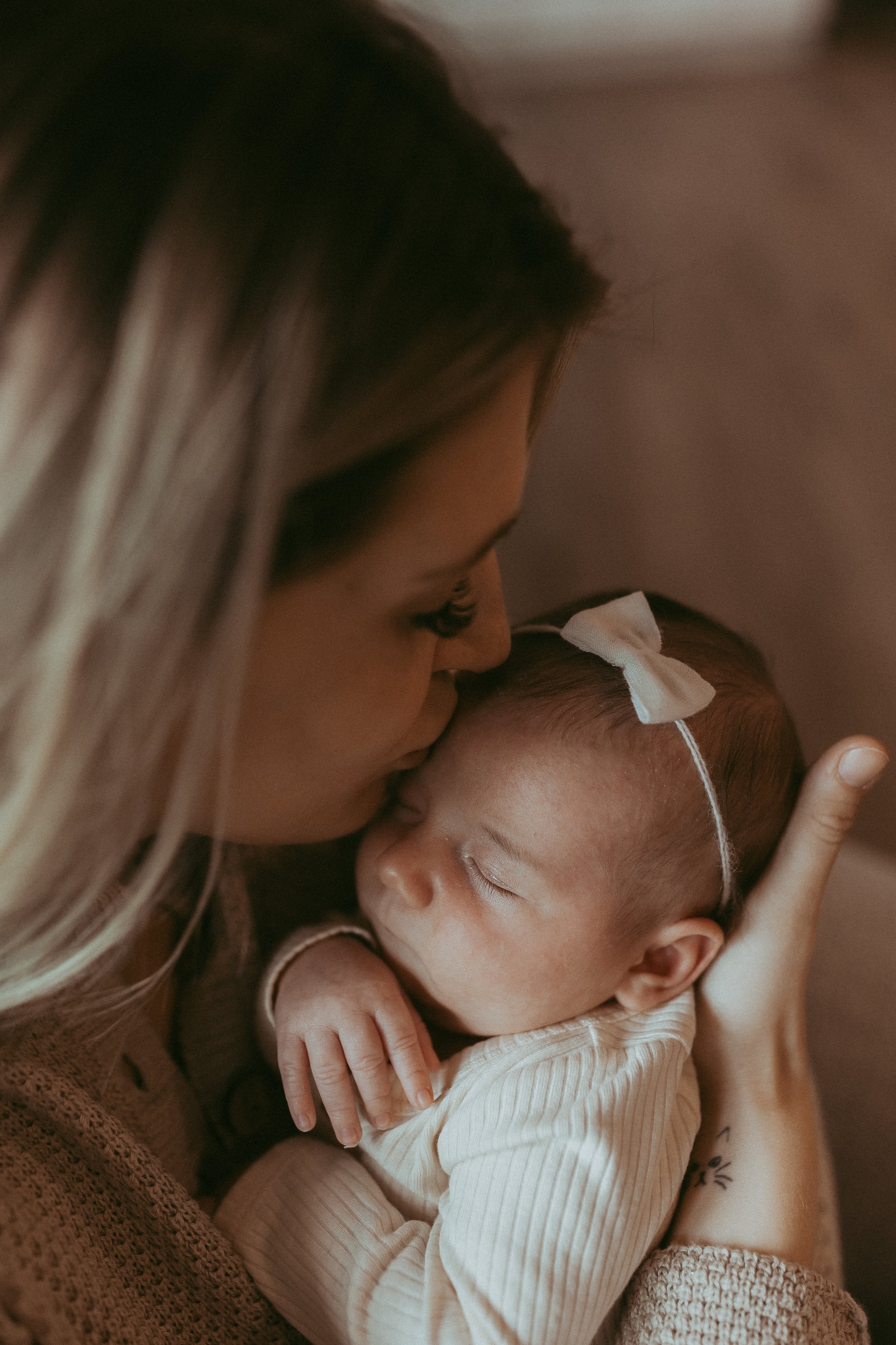A woman is holding and kissing a sleeping newborn baby girl with a bow on her head.
