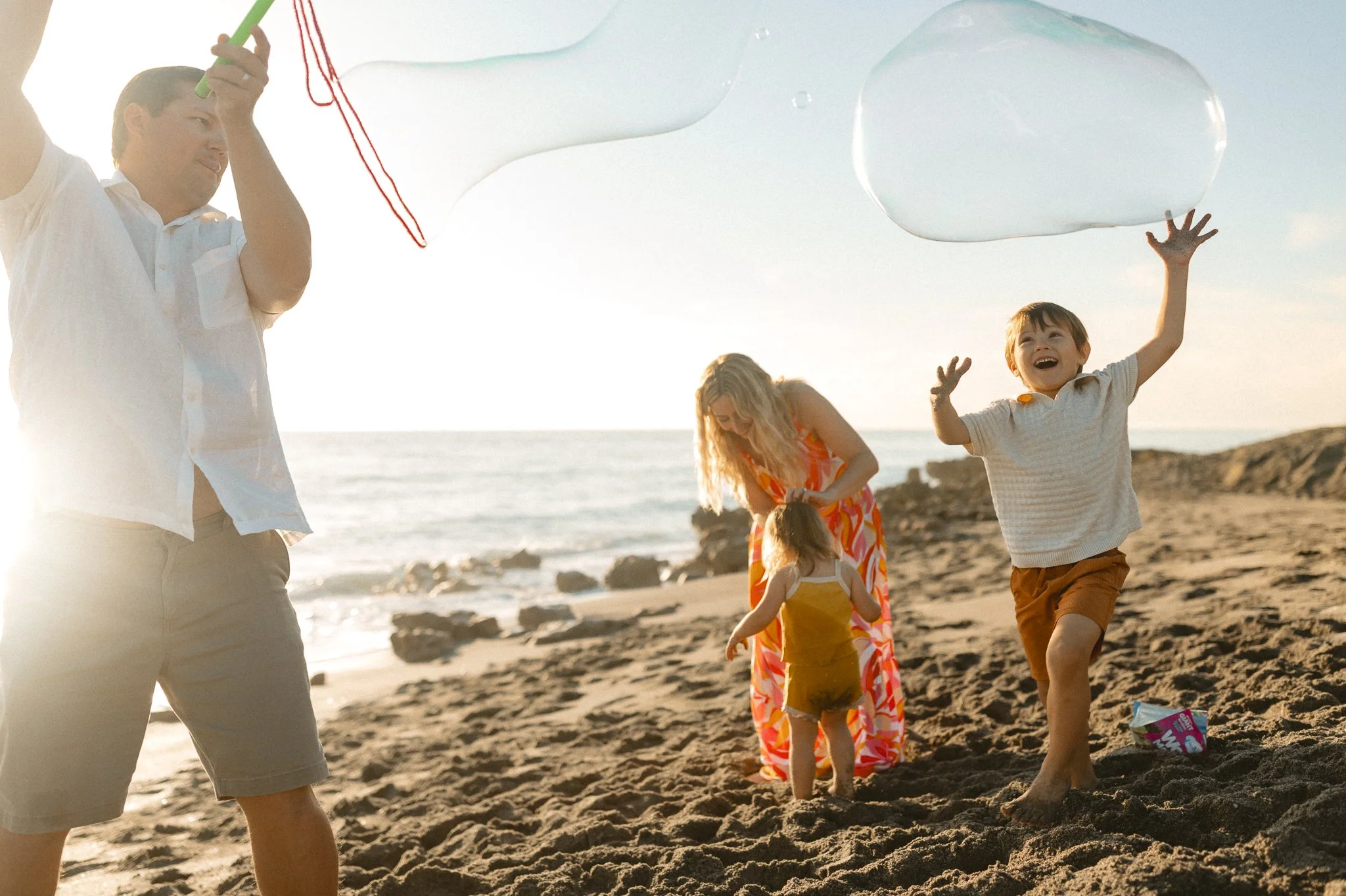 a father blowing big bubbles and his young son jumping and trying to pop it with the mother consoling her young daughter