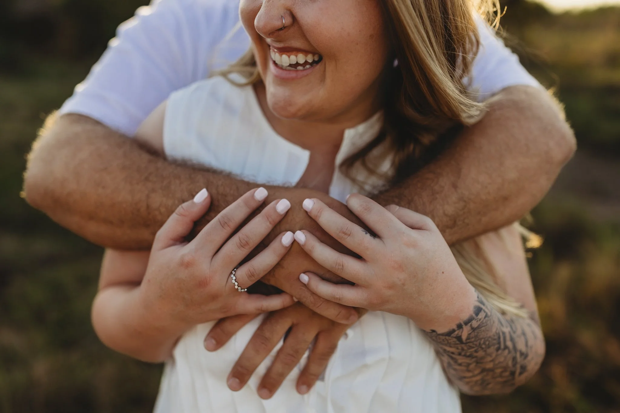A woman with a nose piercing and tattoo on her arm is hugging a man from behind, both smiling. The woman has white nail polish and a ring with small stones on her finger.