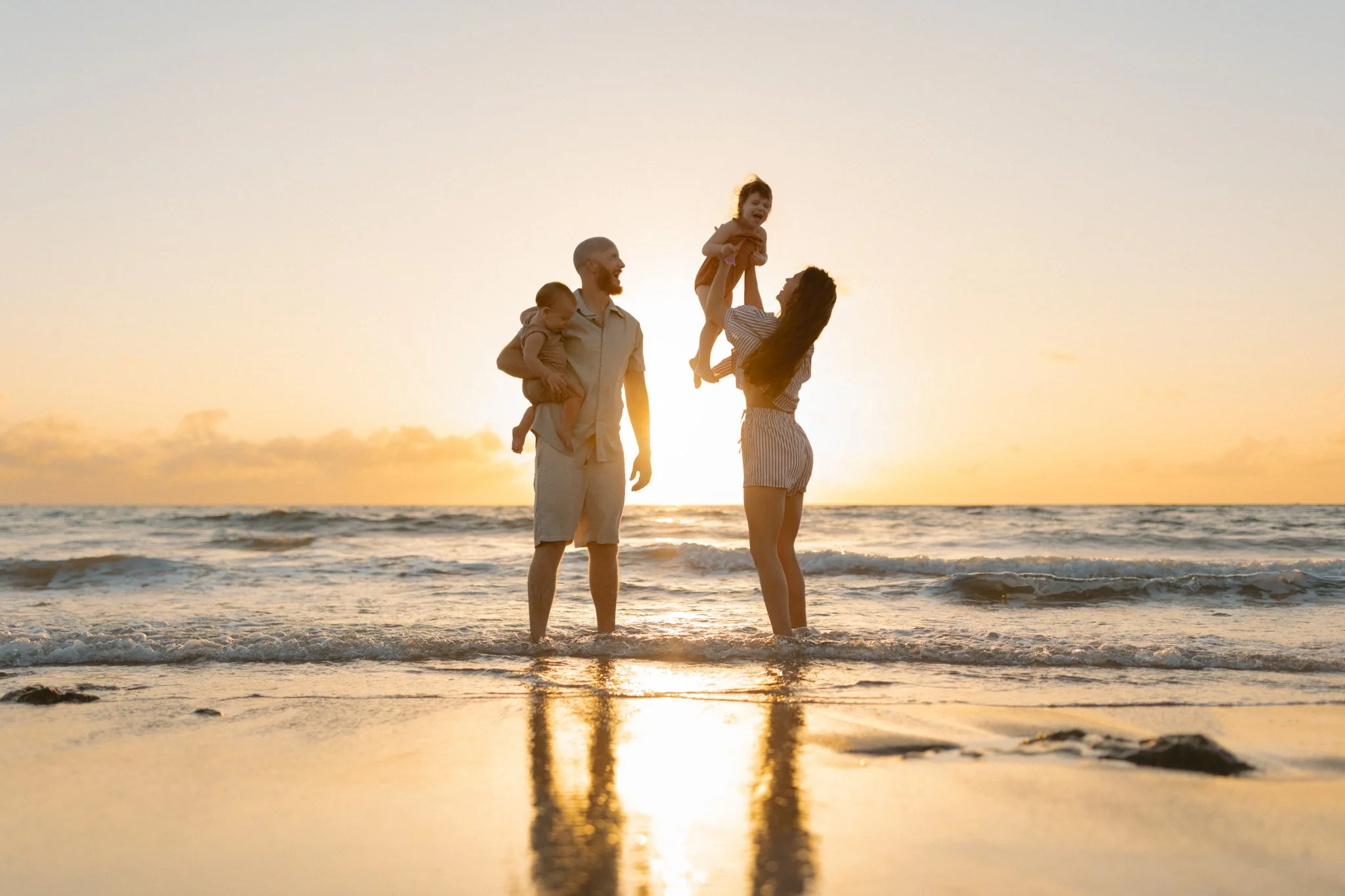 A family of four enjoying a sunset at the beach, with the mother and father holding their two children, one of whom is lifted into the air.