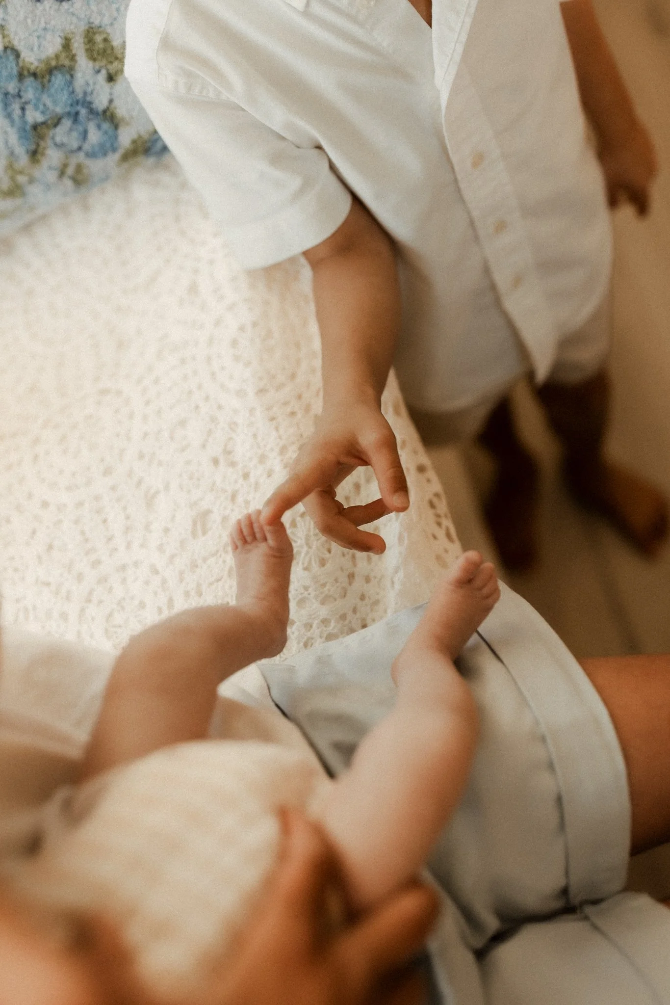 Close-up of an adult holding a baby's tiny hand, both lying on a lace-covered surface in soft, warm lighting.