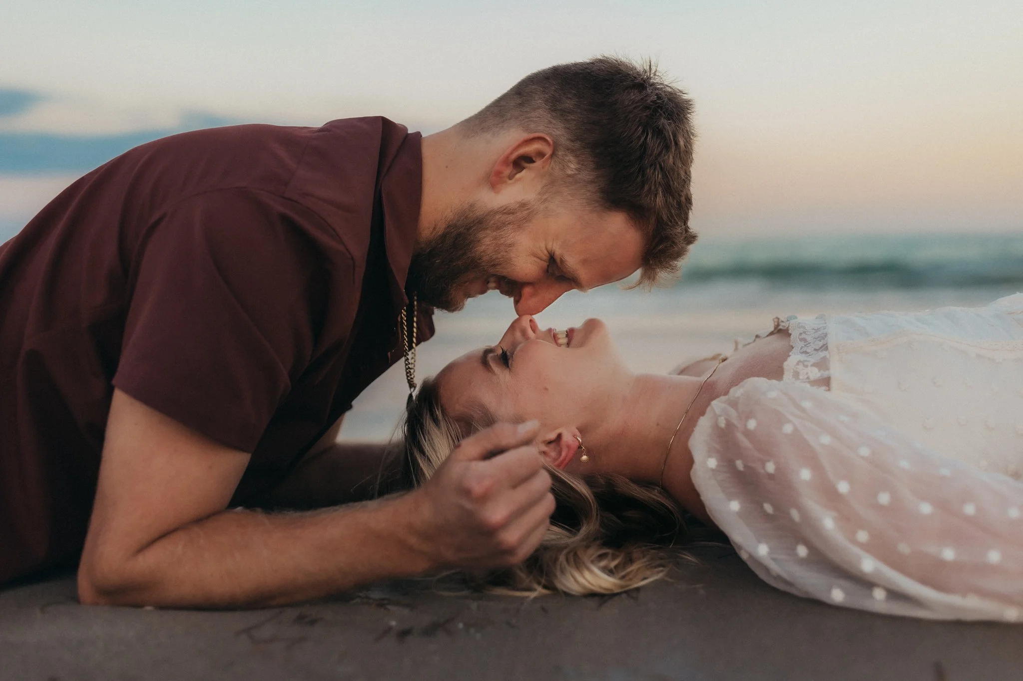 A couple lying on the beach, the man leaning over the woman, both smiling and laughing, with the ocean in the background at sunset.