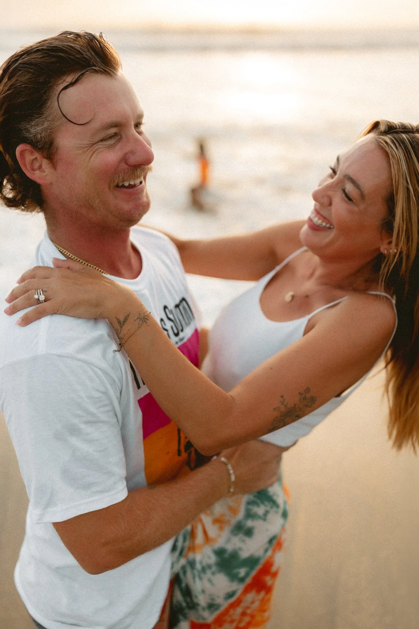 A man and woman smiling and holding each other on a beach at sunset, with the ocean in the background.