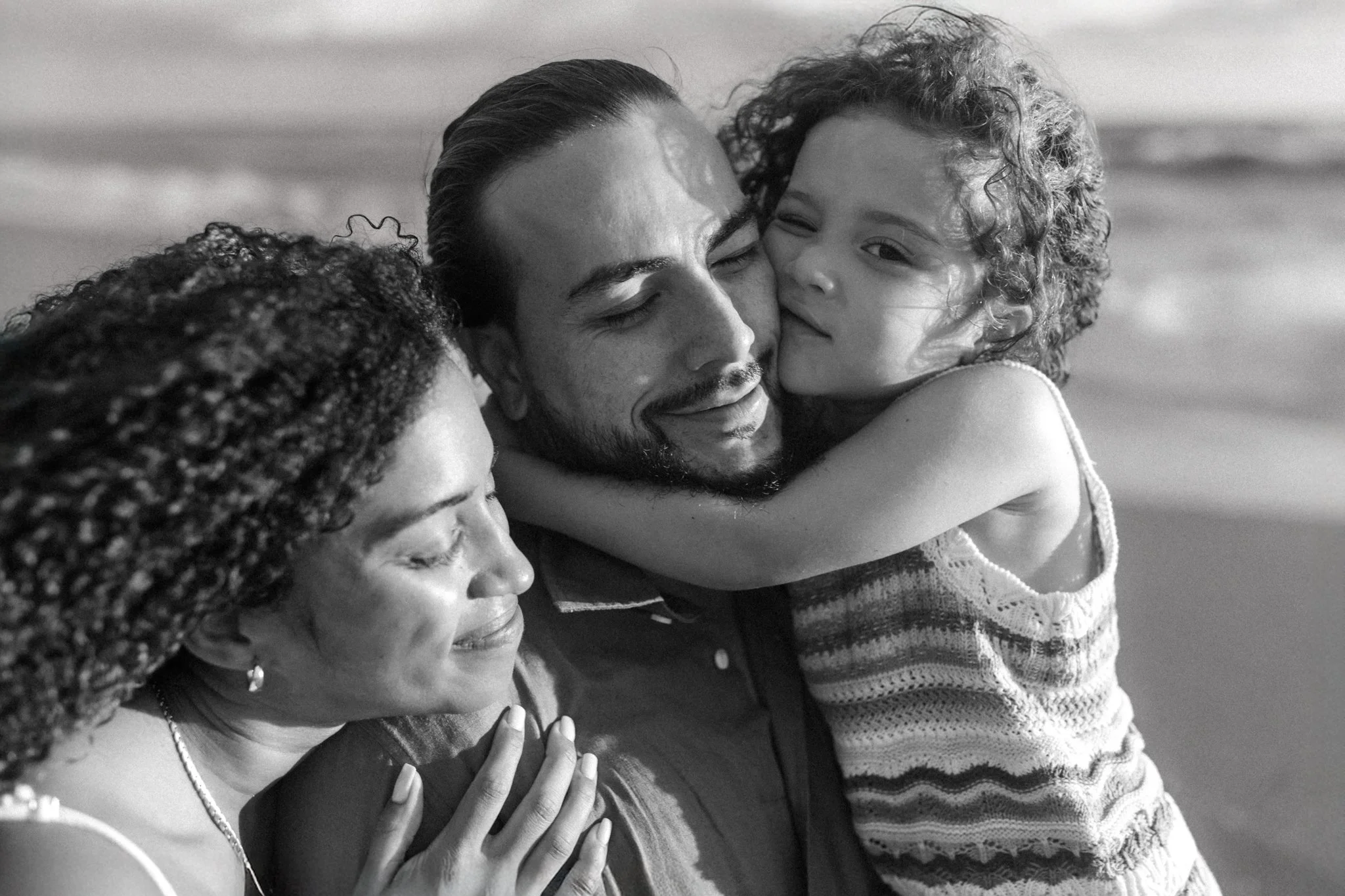 Black and white photo of wife and young child hugging the dad on the beach