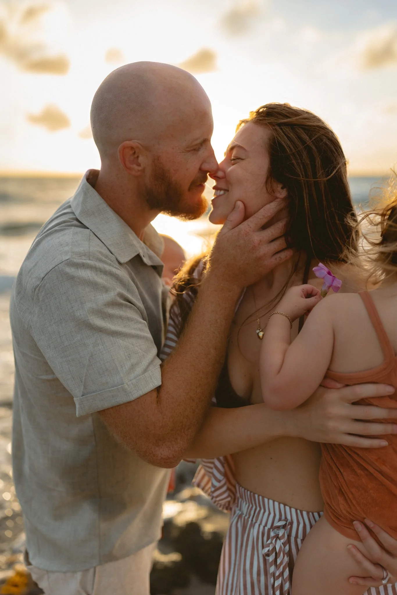 A man and woman touch noses and smile on the beach during sunset, with a woman and child in the scene.