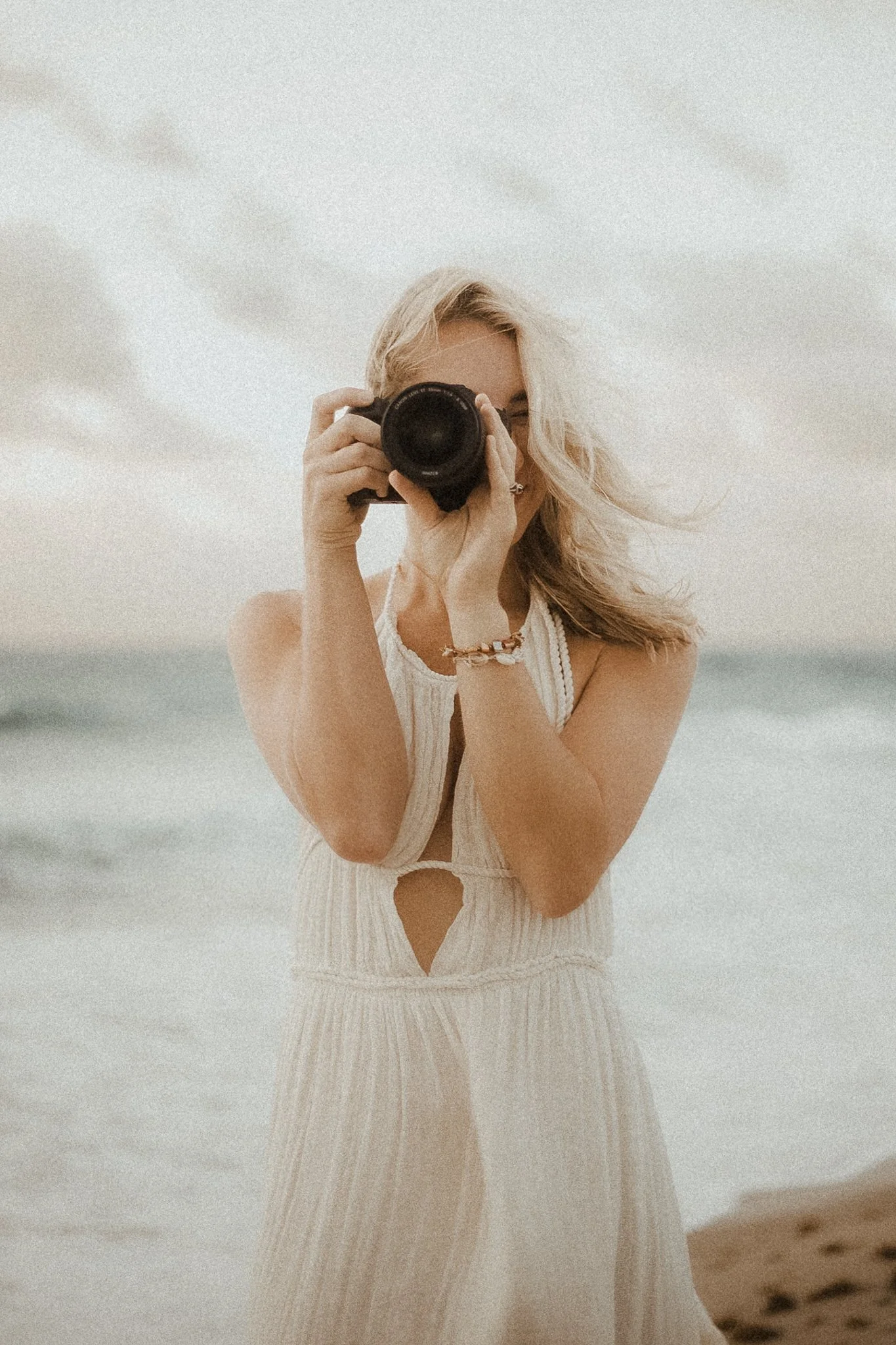 Woman in a white dress taking a photo with a camera at the beach.