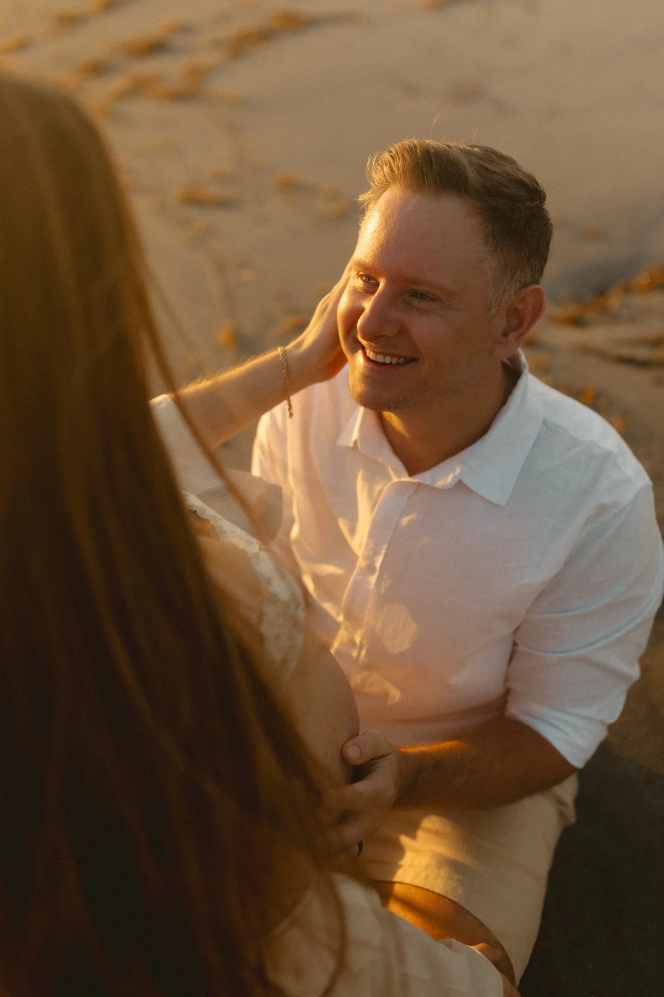 A man kneeling on the beach, smiling at a woman, with the woman touching his face, during sunset.