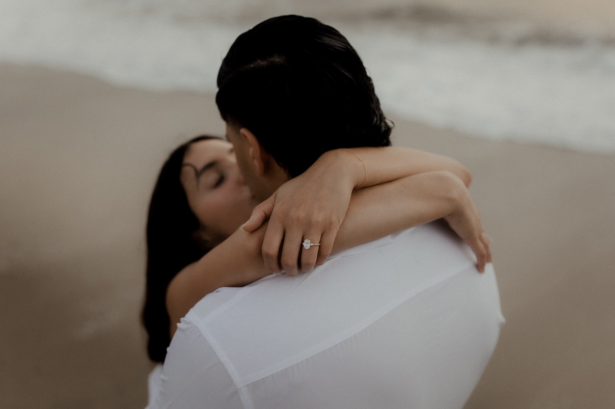 A couple kissing on the beach, with the woman wearing an engagement ring on her finger.