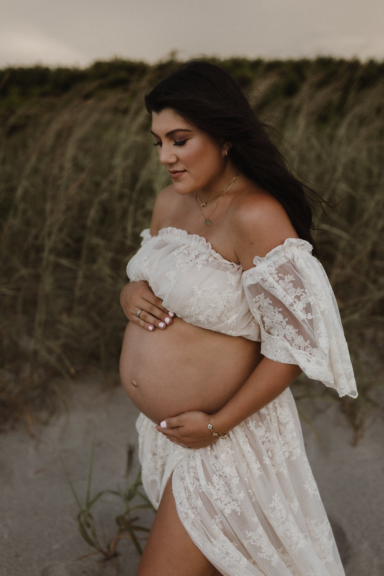 A pregnant woman stands on a sandy beach with tall grass in the background, wearing a white off-the-shoulder lace dress, cradling her belly and smiling softly.