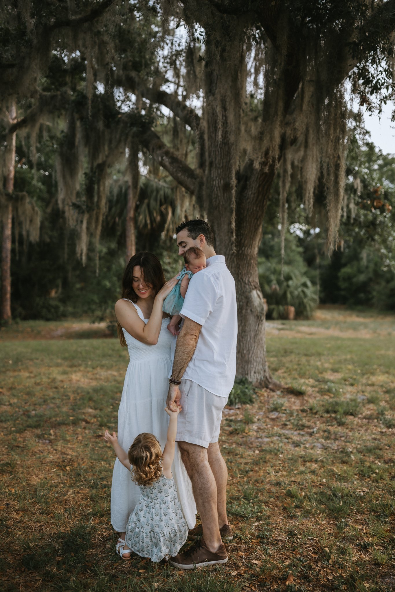 A family of four sharing a joyful moment outdoors in front of a large tree with hanging moss during late afternoon or early evening. The mother, father, a baby, and a young girl are smiling and interacting affectionately.
