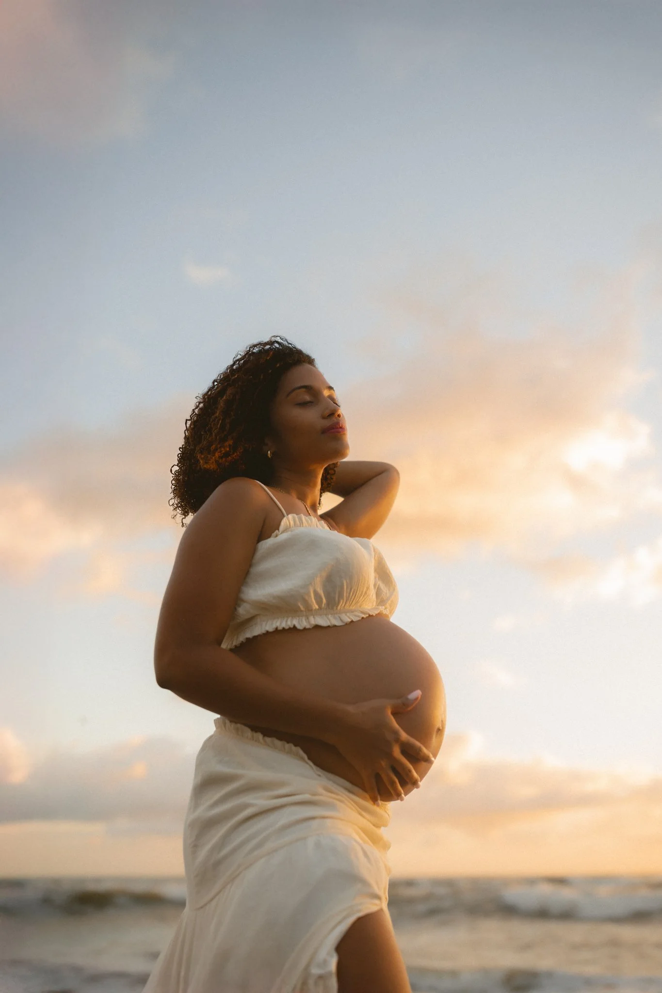 A pregnant woman with curly hair standing on the beach during sunset, wearing a light-colored crop top and skirt, with her eyes closed and one hand on her belly.