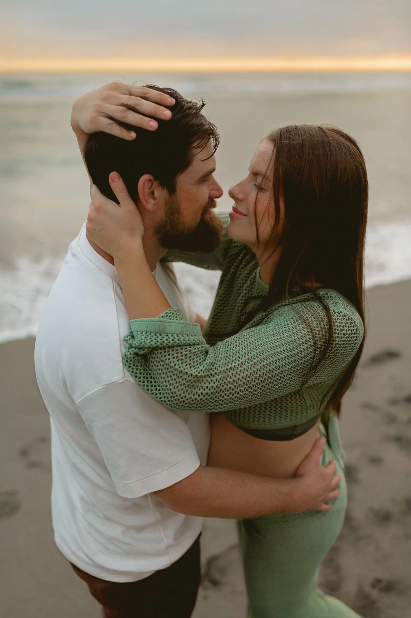 a pregnant lady wearing a green two piece outfit embracing her husband on the beach