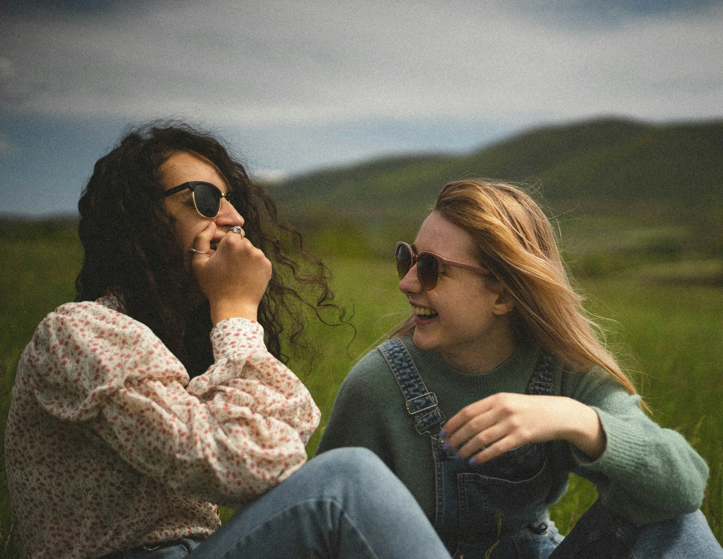 Two women with sunglasses smiling and laughing outdoors in a green field, facing each other close up. queer couple, lesbian couple, queer therapy, EFT, emotion focused therapy.