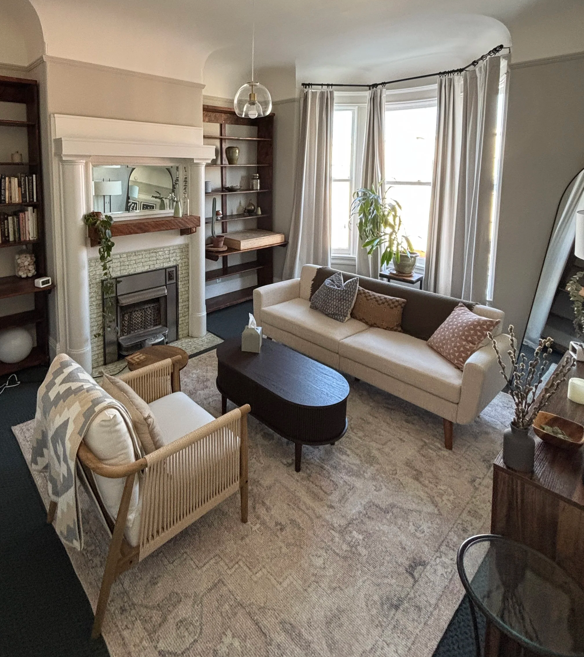 Living room with beige sofa, wooden coffee table, armchair, fireplace with white mantel, built-in wooden shelves, large window with white curtains, potted plant, patterned area rug, and a decorative floor lamp.