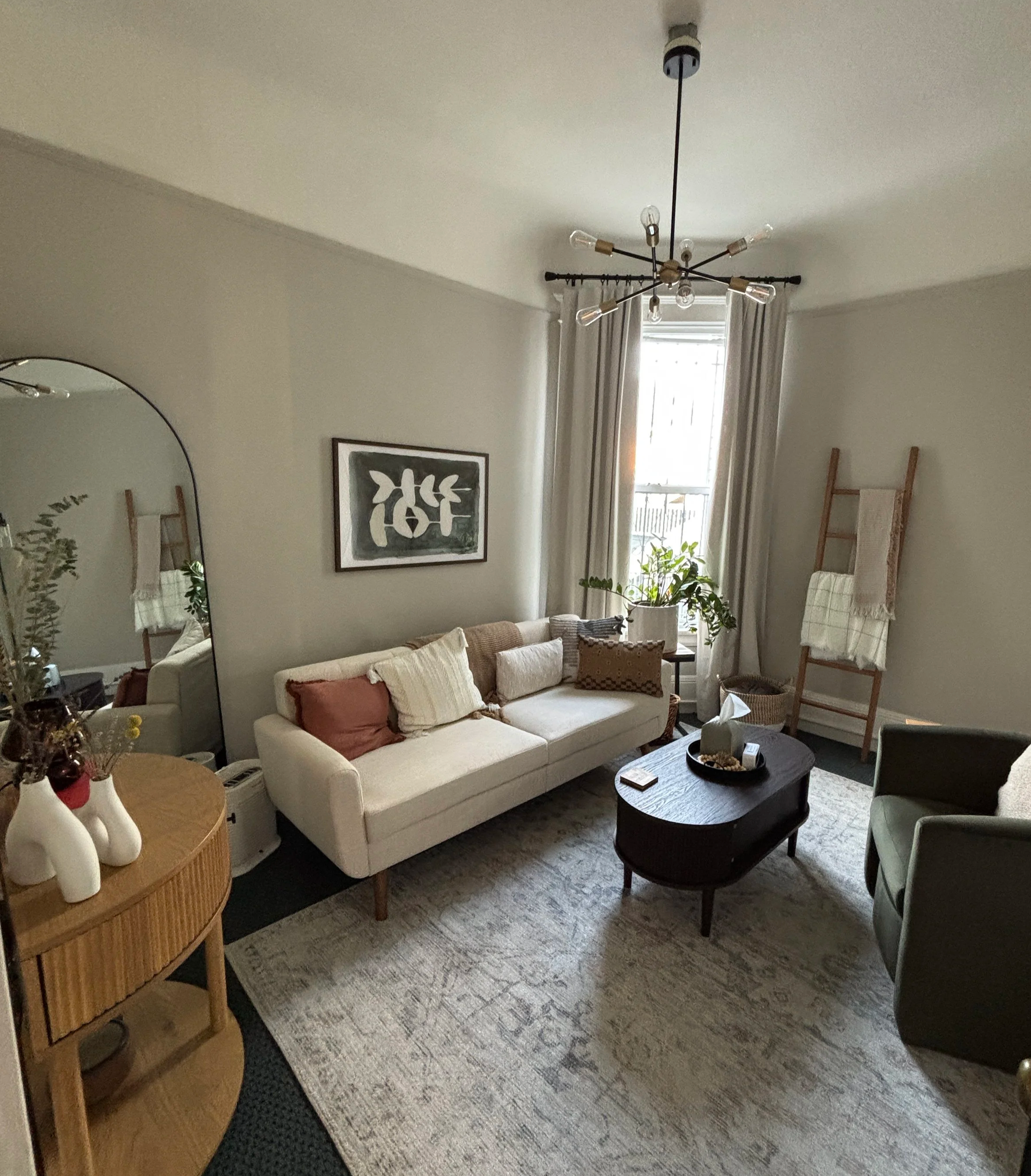 A cozy living room with a white sofa decorated with various pillows, a wooden coffee table with decorative items, a patterned rug, a tall ladder-style shelf with blankets, a framed abstract art piece, a vintage-style chandelier, a window with curtains, and a potted plant.