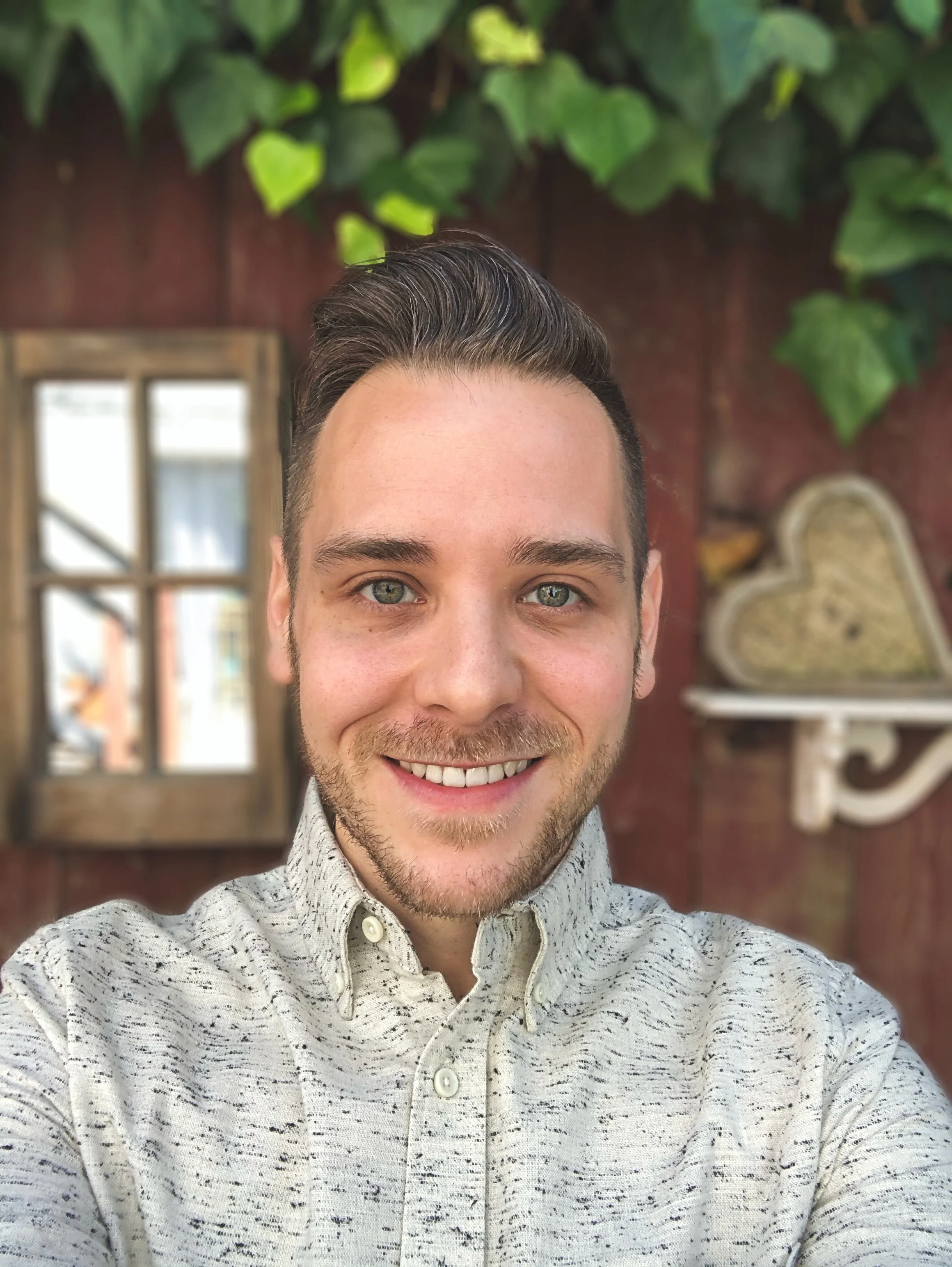 A young man smiling, wearing a speckled white shirt, in an indoor setting with plants and a window in the background.