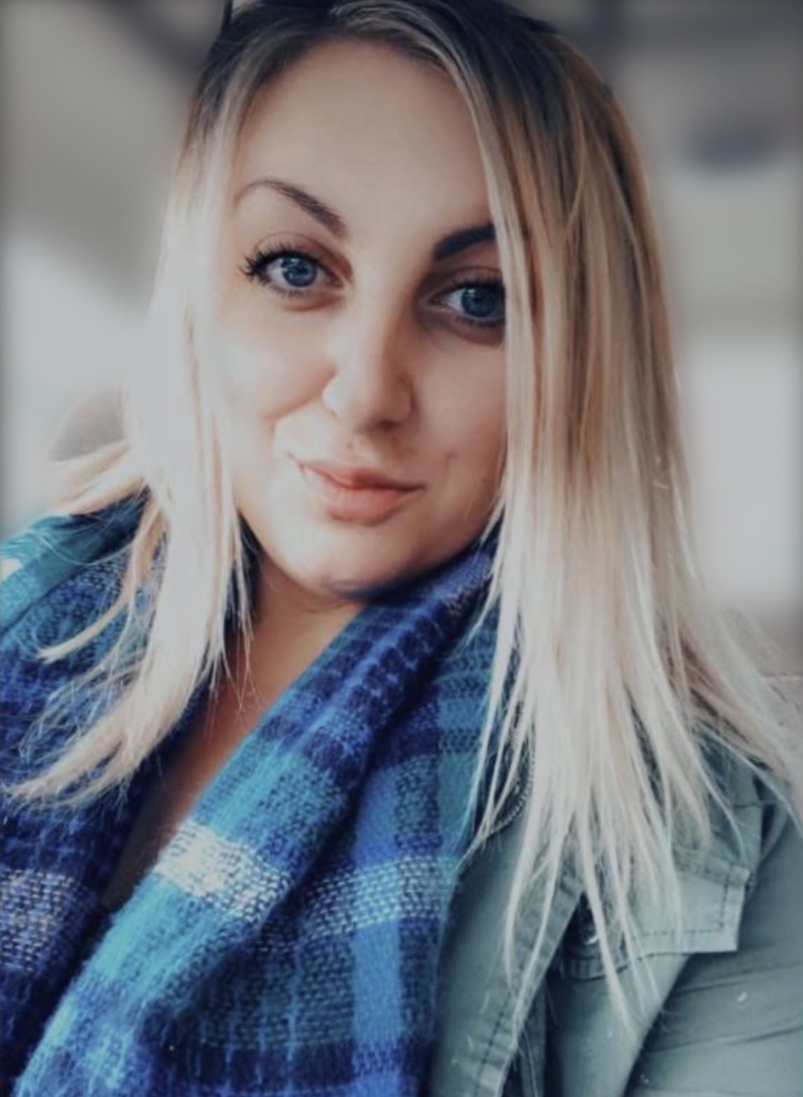 Close-up of a woman with blue eyes and blonde hair, wearing a blue checked scarf and a green jacket.