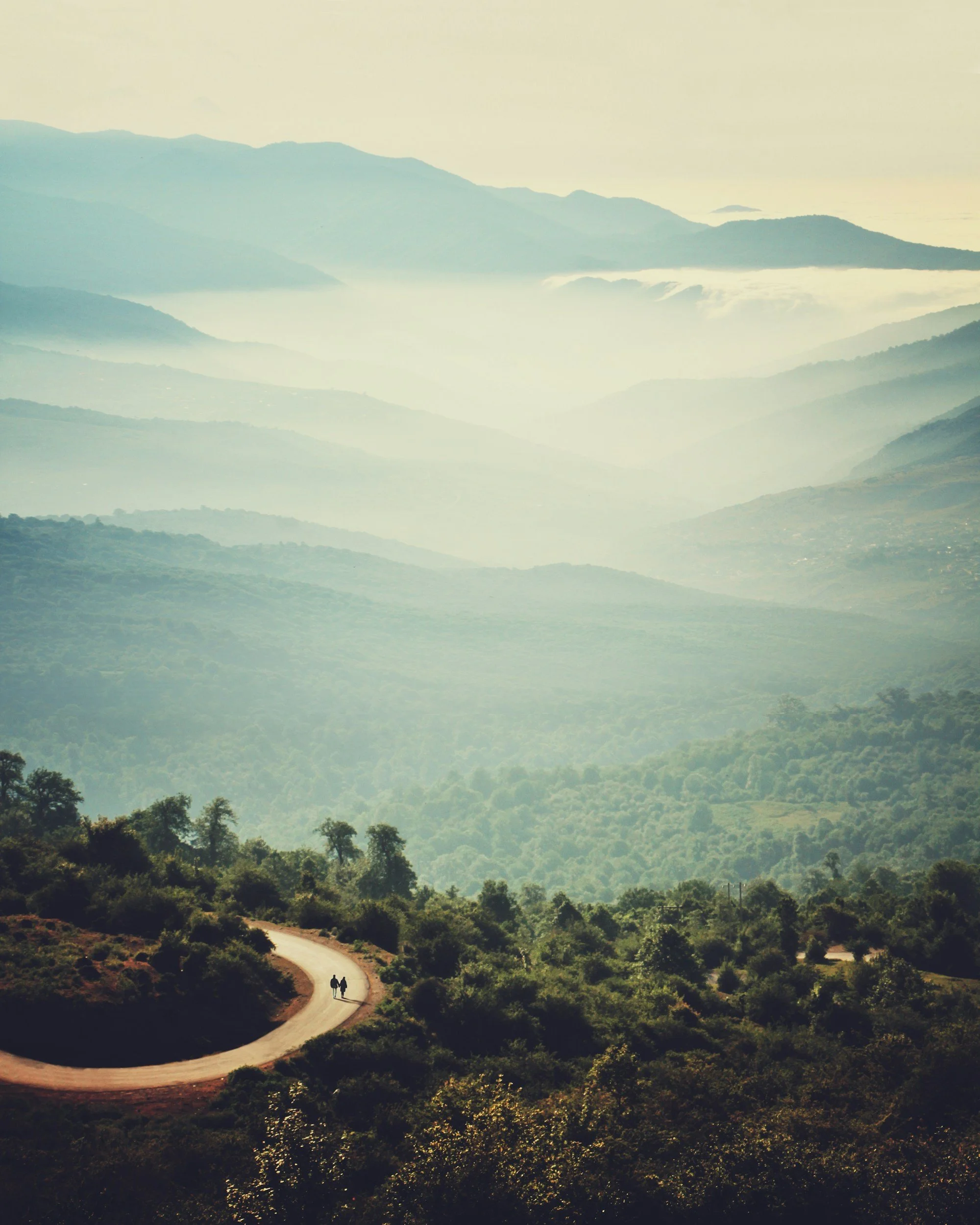 A winding dirt road through green forested hills with distant mountain ranges and a pale sky.