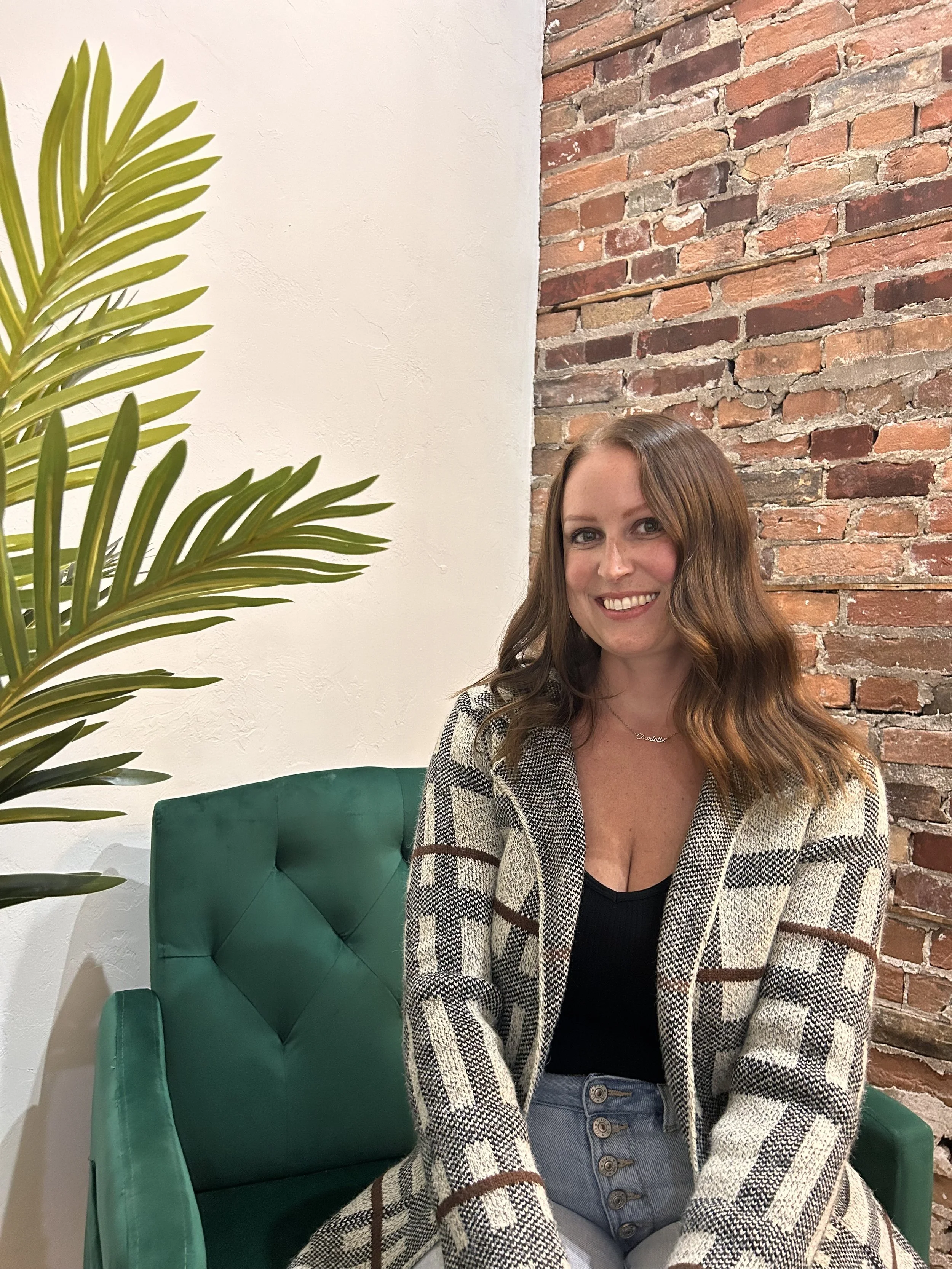 A woman with wavy brown hair smiling, wearing a black top and plaid blazer, sitting in a green armchair next to a tall plant with large green leaves, in front of a brick wall and white wall.