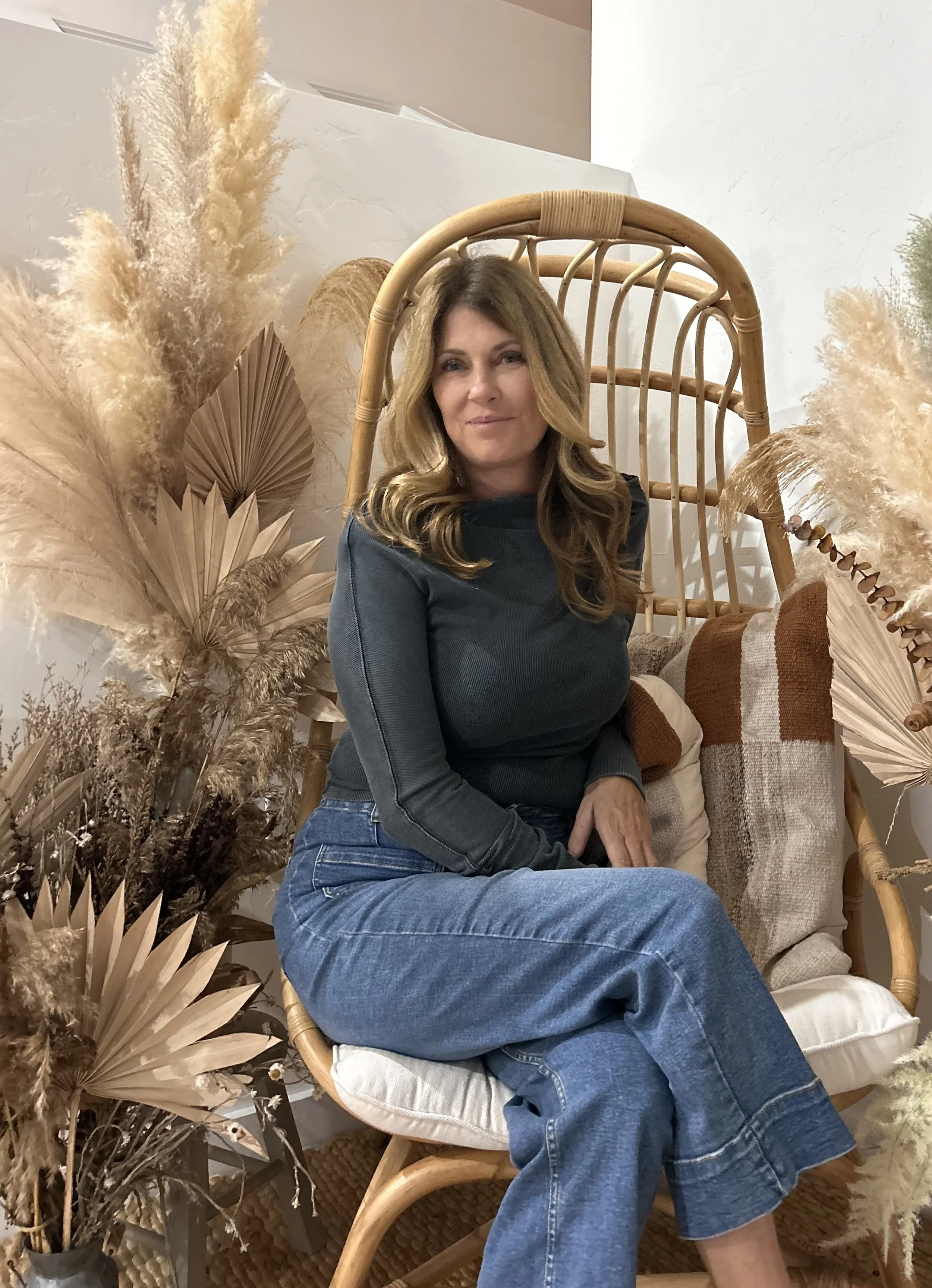 Vanessa Blanchette, wearing a dark long-sleeve shirt and blue jeans, sitting on a beige cushion in a wicker hanging chair surrounded by beige and brown dried decorative plants and leaves, in an interior setting with neutral-colored walls.