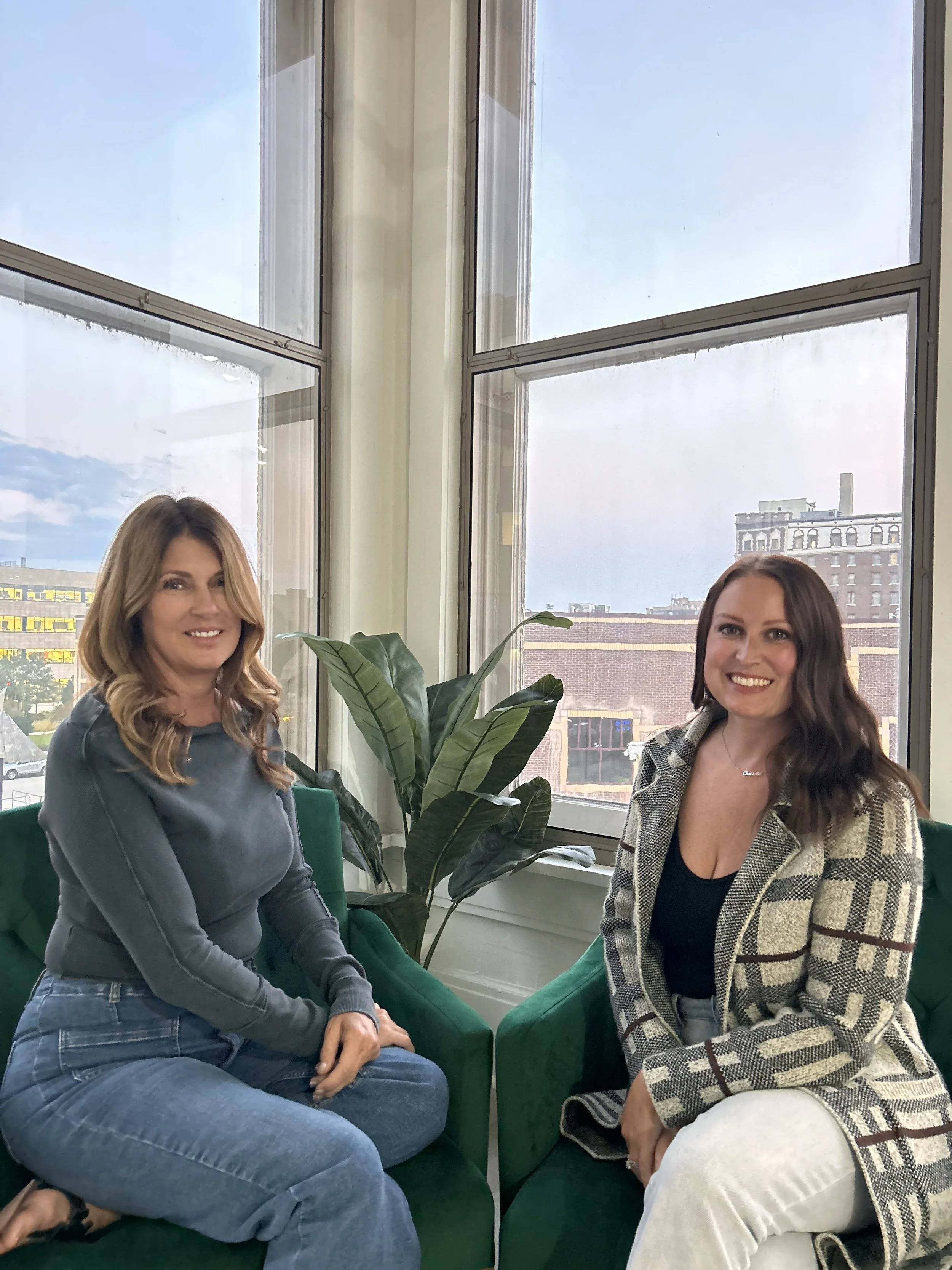 Two women sitting on green chairs in a bright room with large windows, city buildings outside, and a large potted plant between them.