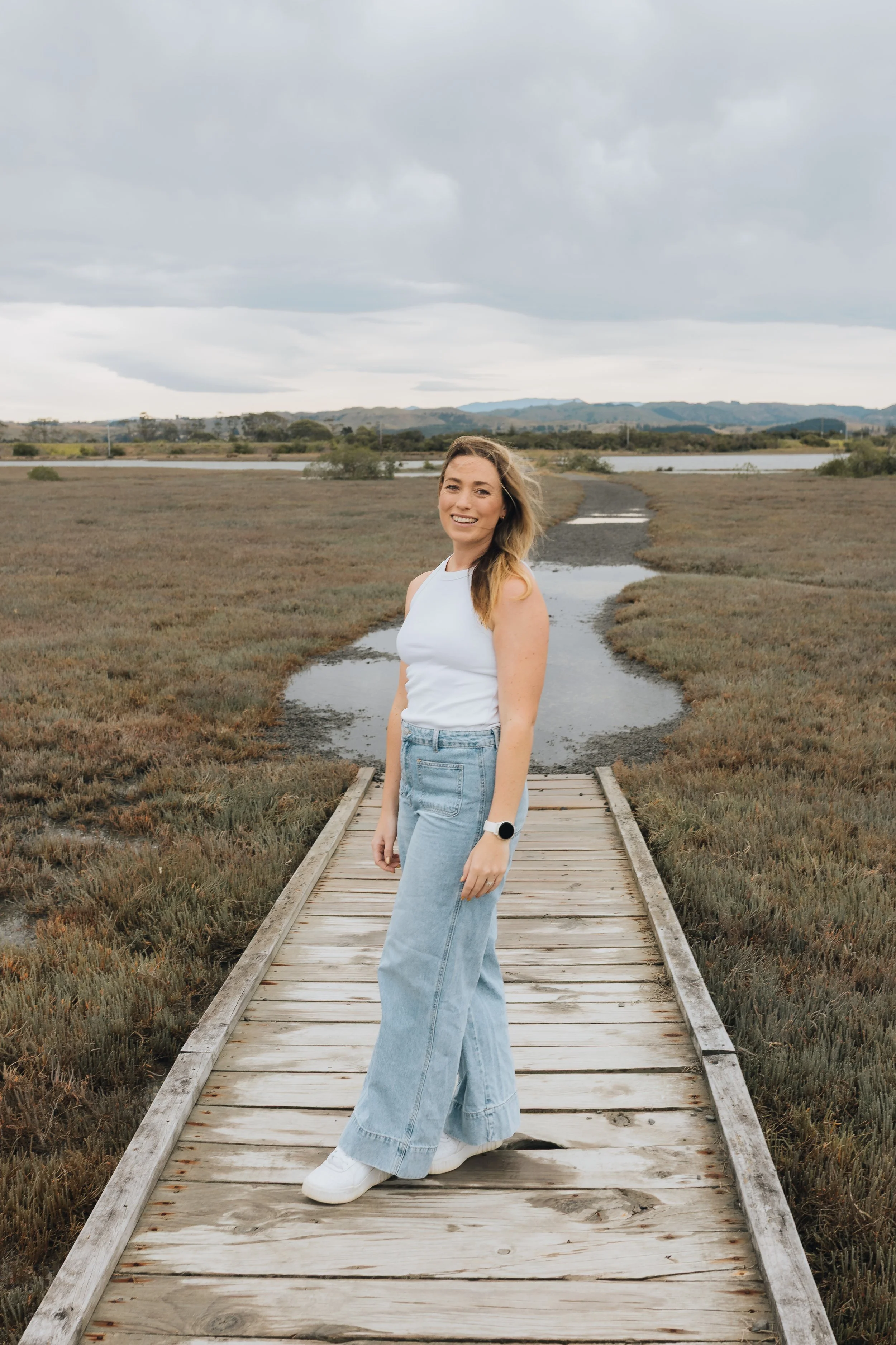 A woman with long hair smiling, standing on a small wooden bridge in a marshy wetland area with water, grasses, and distant rolling hills under a cloudy sky.