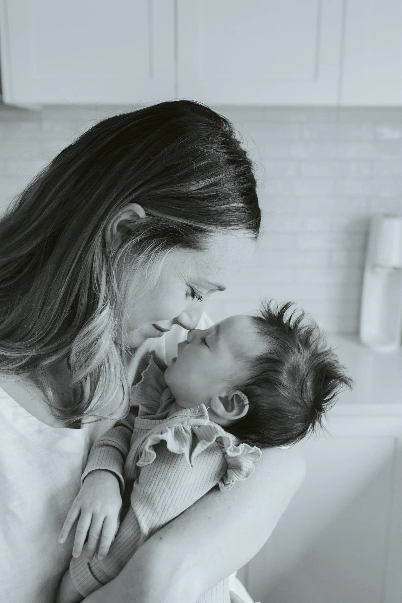 A woman and a young girl sharing a close moment with their foreheads touching, both with their eyes closed, in a room with light-colored walls and a white cabinet in the background.