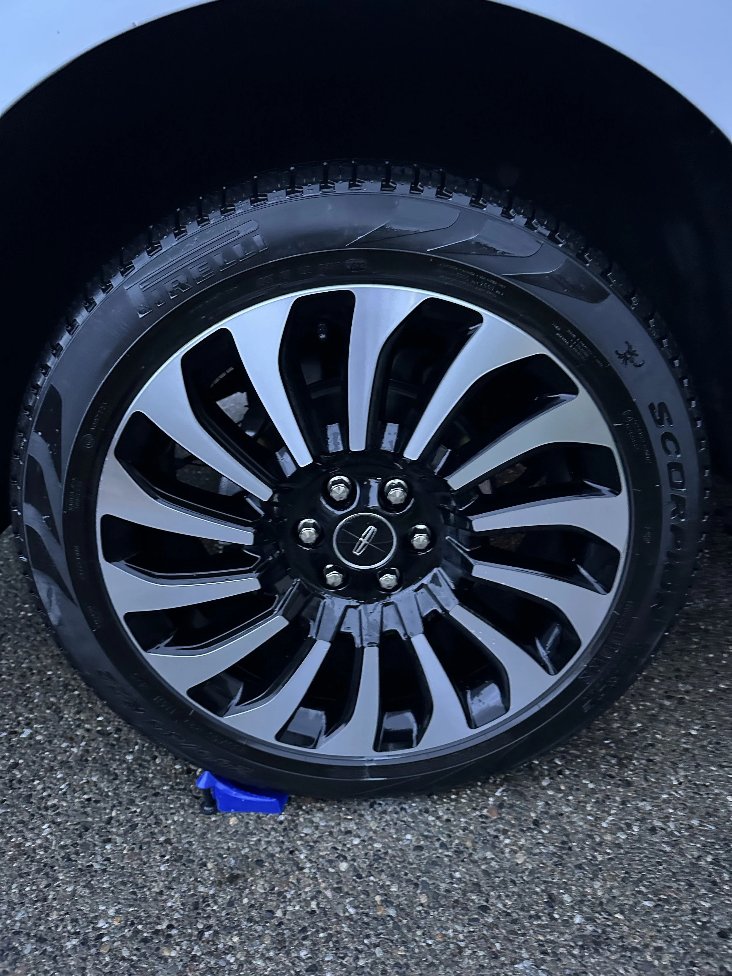 Close-up of a car tire and alloy wheel on asphalt, with a small blue object near the bottom of the tire. After the clean