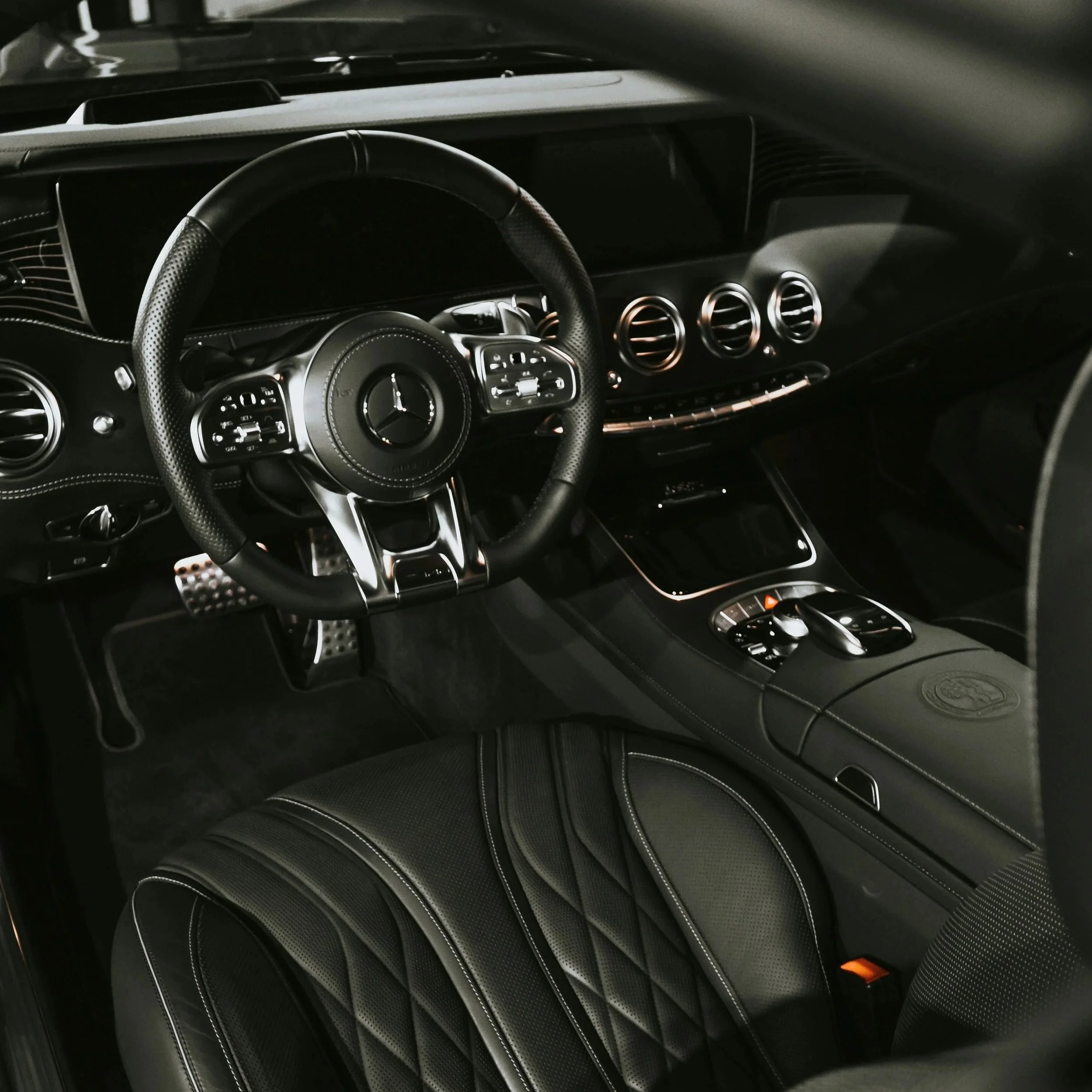 Interior of a luxury car, showing a steering wheel with Mercedes-Benz logo, dashboard with air vents, gear shift, and black leather seat with detailed stitching.