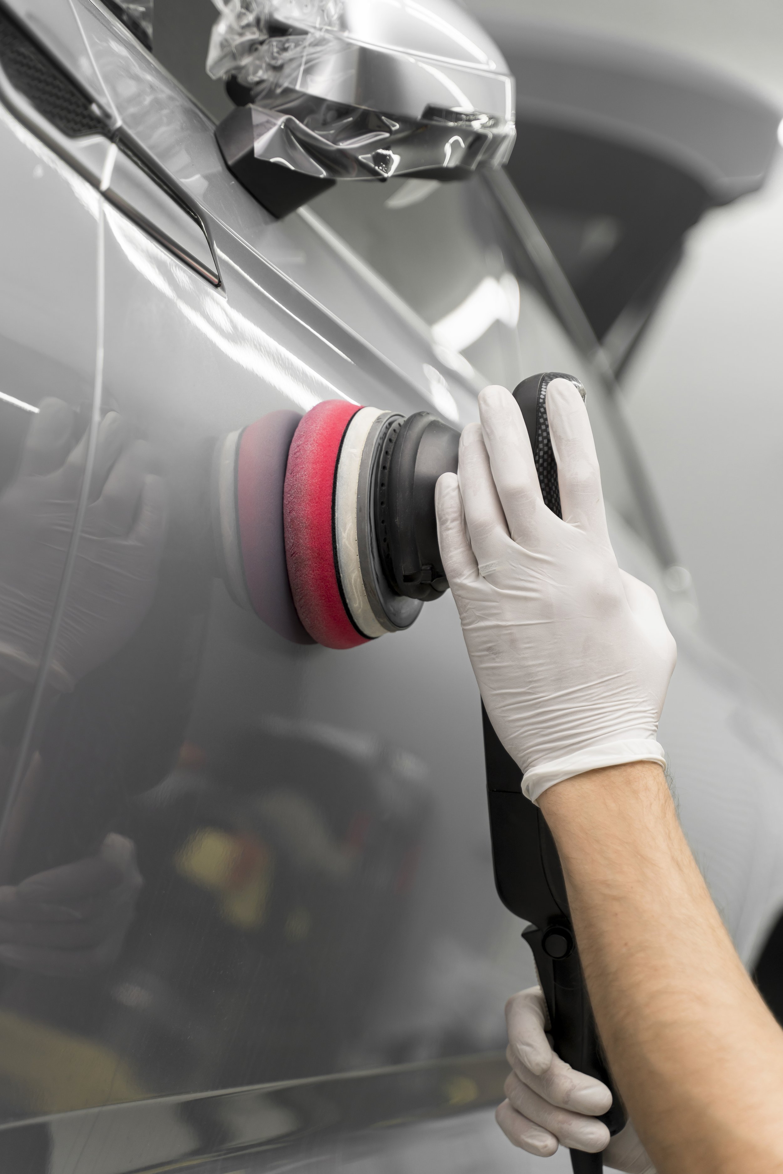 A person wearing white gloves polishes a gray car's exterior using a handheld polishing machine with a red and black pad.