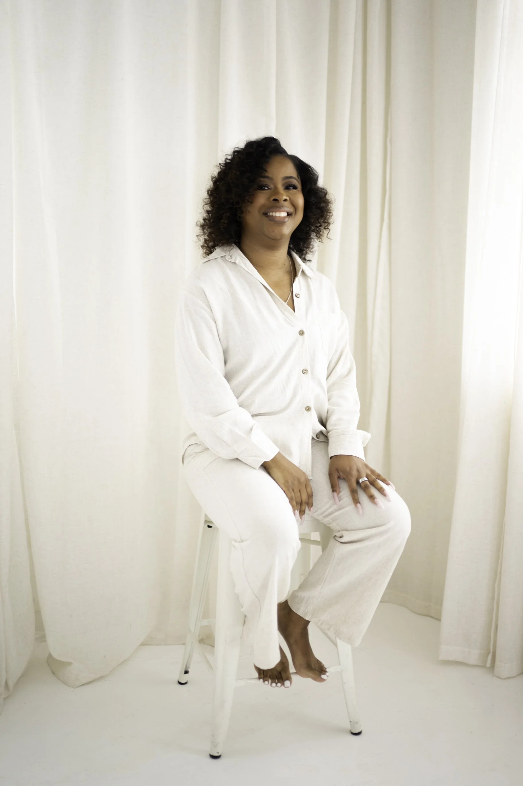 A woman with curly hair smiling, sitting on a white chair in front of white curtains, dressed in all white, with her hand resting on her knee.