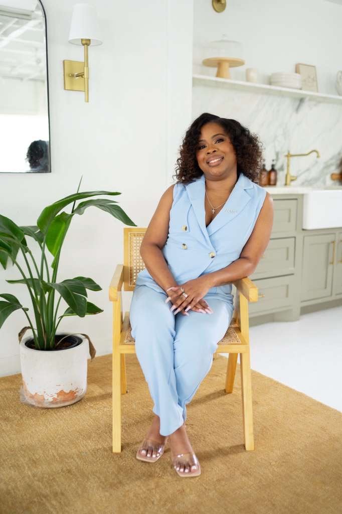A woman with curly hair sitting on a wooden chair in a modern kitchen, wearing a light blue sleeveless pantsuit, smiling, with a potted plant nearby and a mirror on the wall.