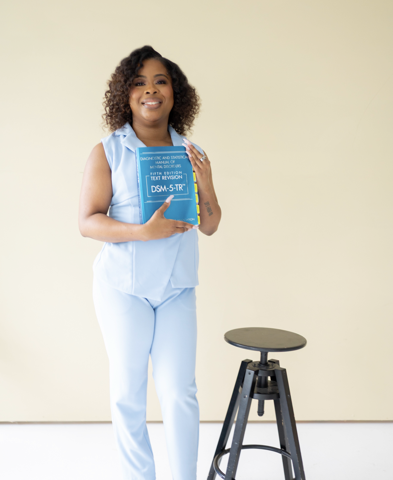 A woman with curly hair in a light blue outfit holding a DSM-5 manual, standing next to a stool against a plain beige background.