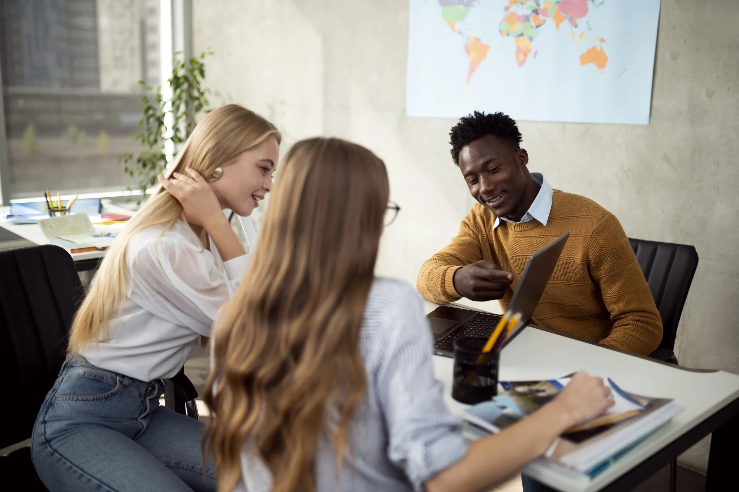 Group of four young adults sitting around a table in a modern office, engaged in a discussion. A world map is on the wall behind them, and office supplies are visible on the table and windowsill.