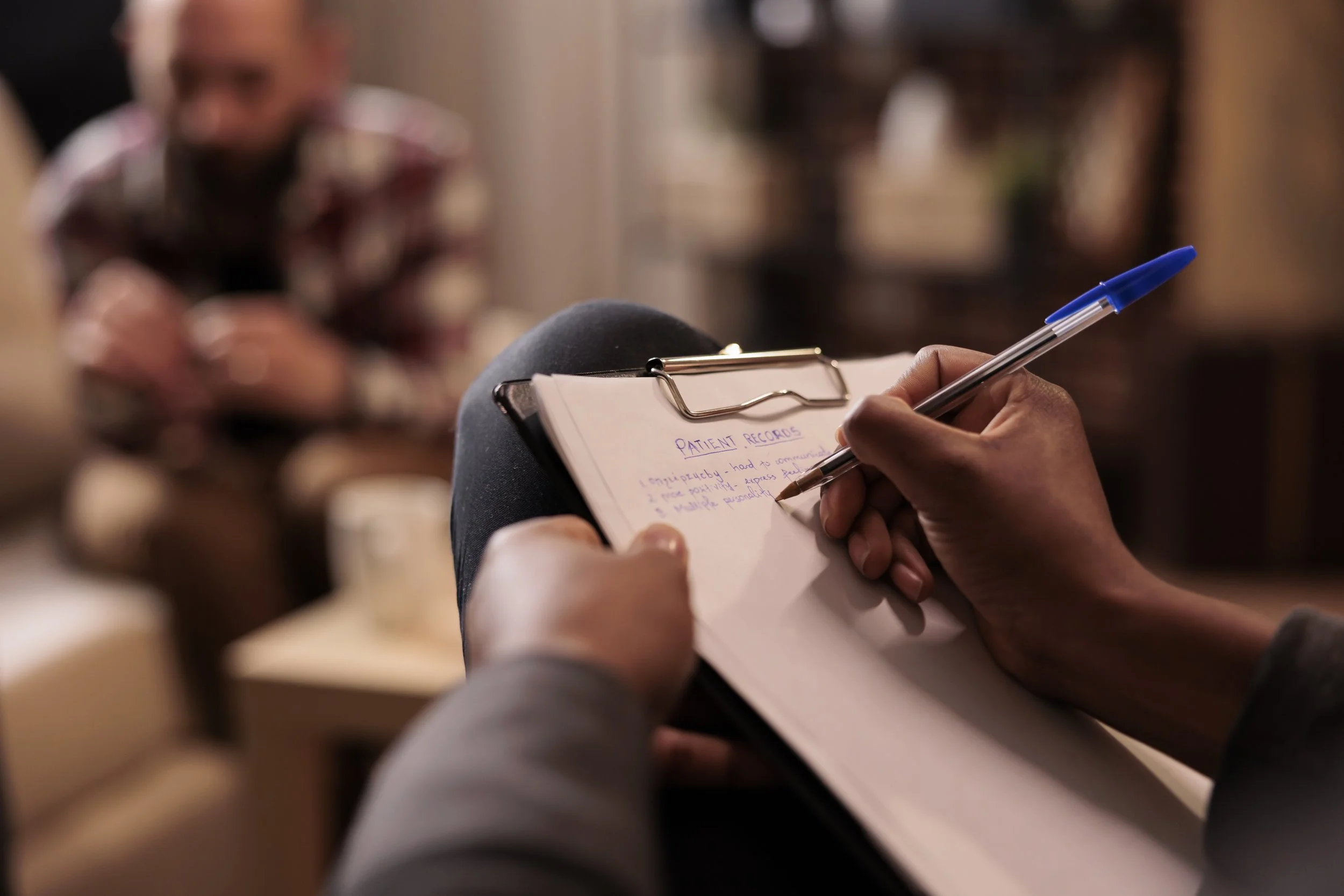 A person holding a pen and taking notes on a clipboard with a label that says 'PATIENT RECORDS' in a setting with blurred figures in the background.