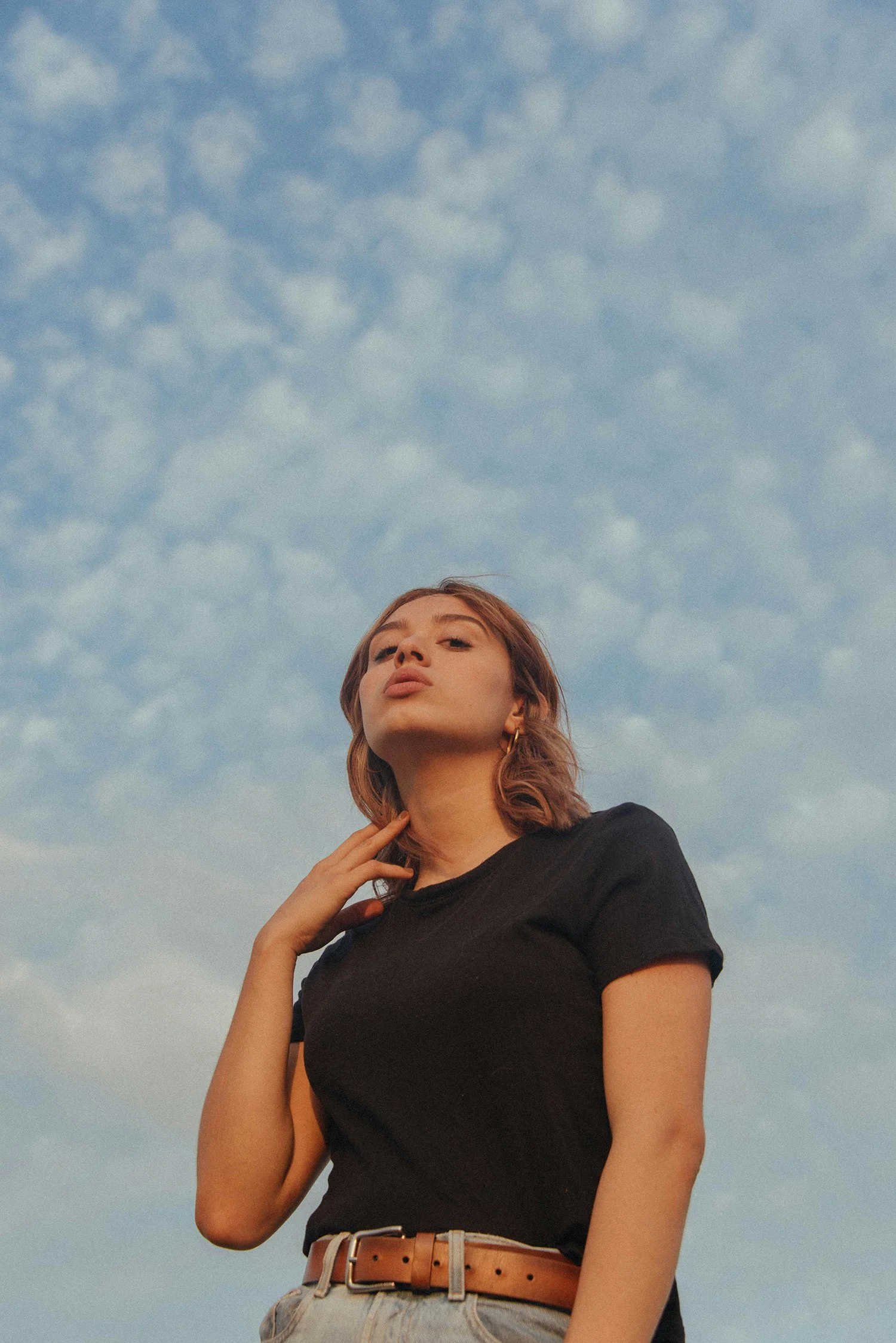 En ung kvinna står under en molnig himmel, bär en svart t-shirt och ljusa jeans, med handen mot halsen och tittar in i kameran.