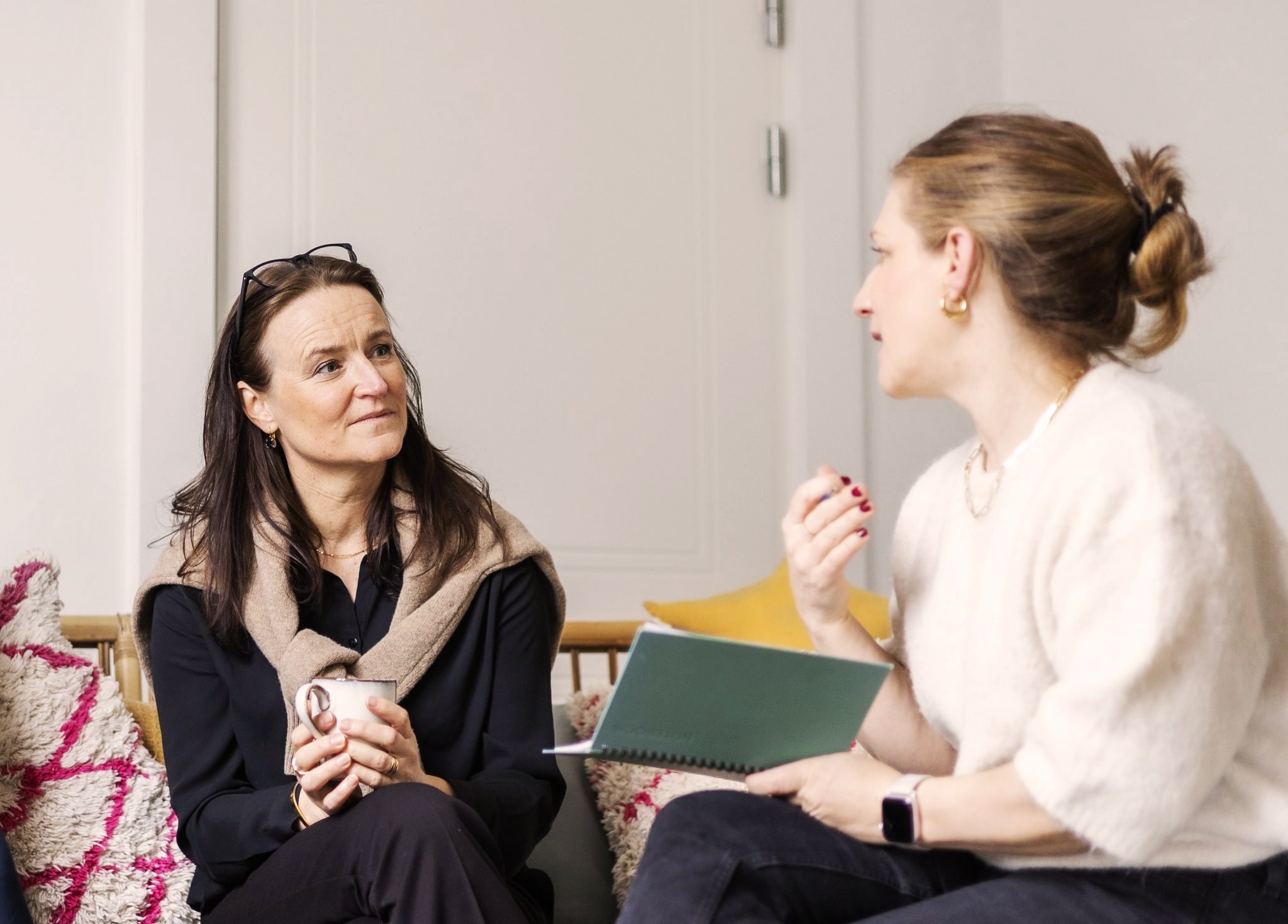 Två kvinnor ur personalen på Stella pratar i ett rum. Den vänstra kvinnan håller en mugg, den högra kvinnan håller en öppen bok eller anteckningsblock.