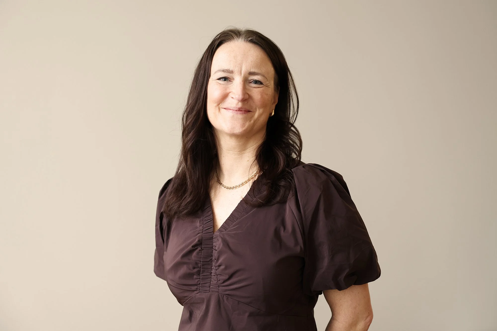 Personal på Stella Barn & Ungdom med mörkt, vågigt hår, iklädd en mörkbrun blus, står mot en beige bakgrund och ler.