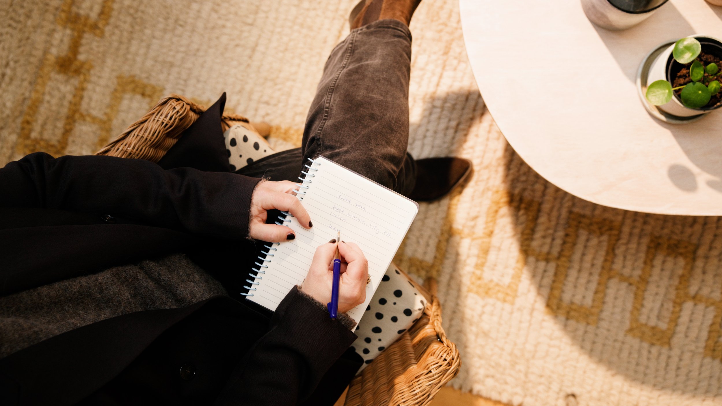 Terapeut som sitter i en stol, skriver i en anteckningsbok, med en kopp och en växt på ett runt bord.