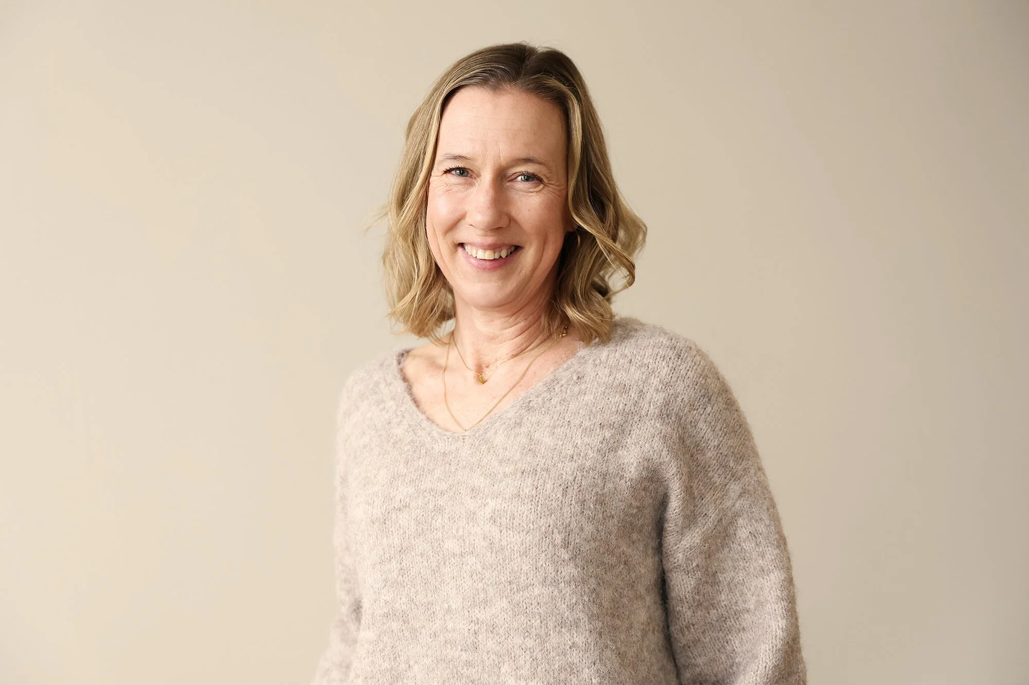 Personal på Stella Barn & Ungdom med axellångt blont hår, iklädd en beige stickad tröja, står framför en ljus beige bakgrund.