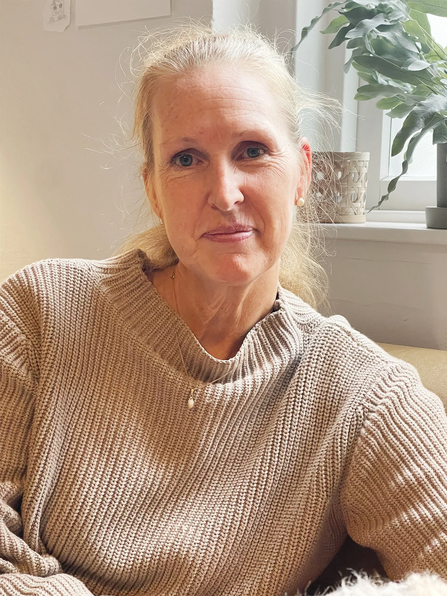 Personal på Stella Barn & Ungdom med ljusblont hår, iförd en beige stickad tröja, sitter i ett rum med växter och en fönster i bakgrunden.
